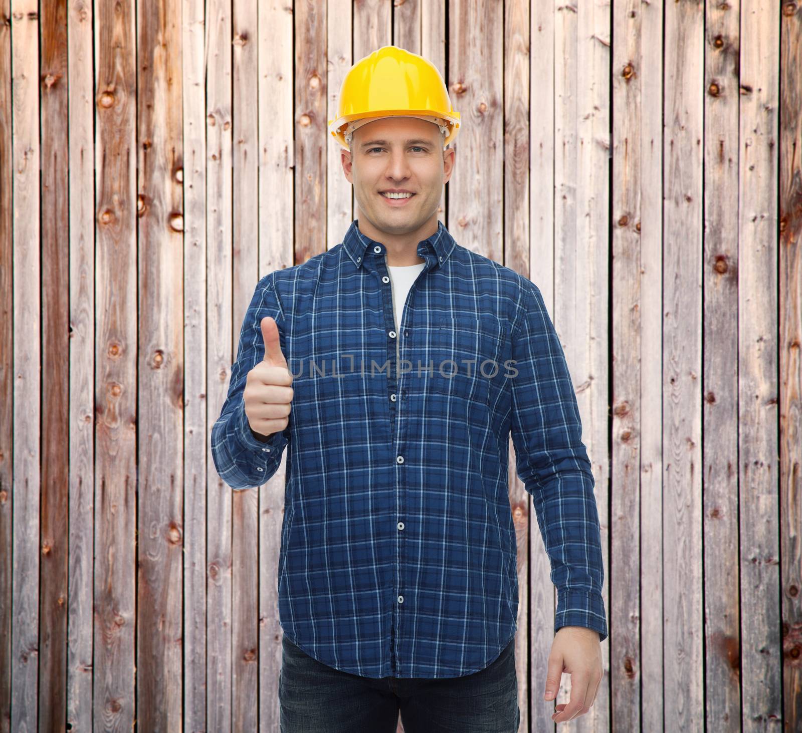 repair, construction, building, people and maintenance concept - smiling male builder or manual worker in helmet showing thumbs up over wooden fence background