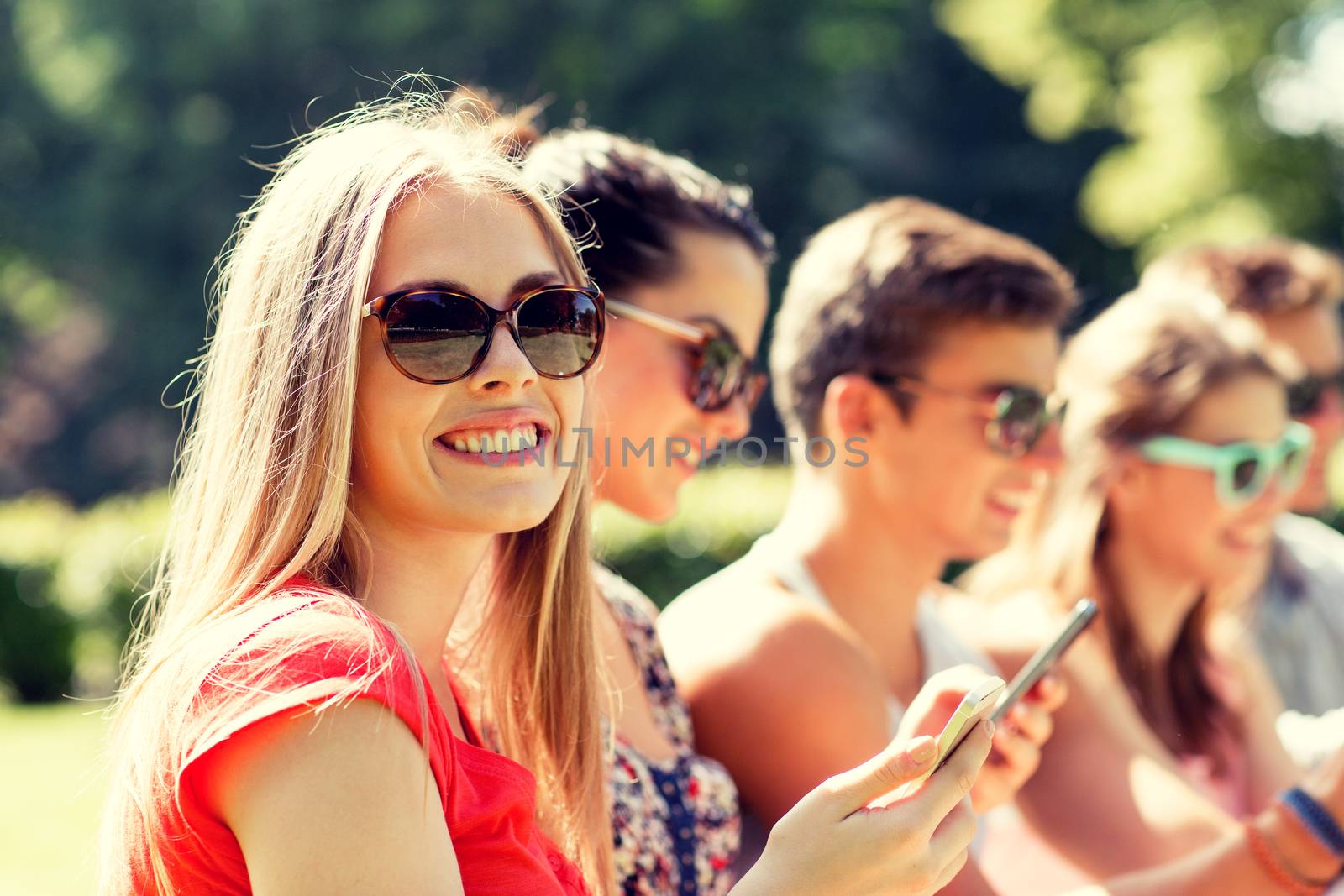 smiling friends with smartphones sitting in park by dolgachov