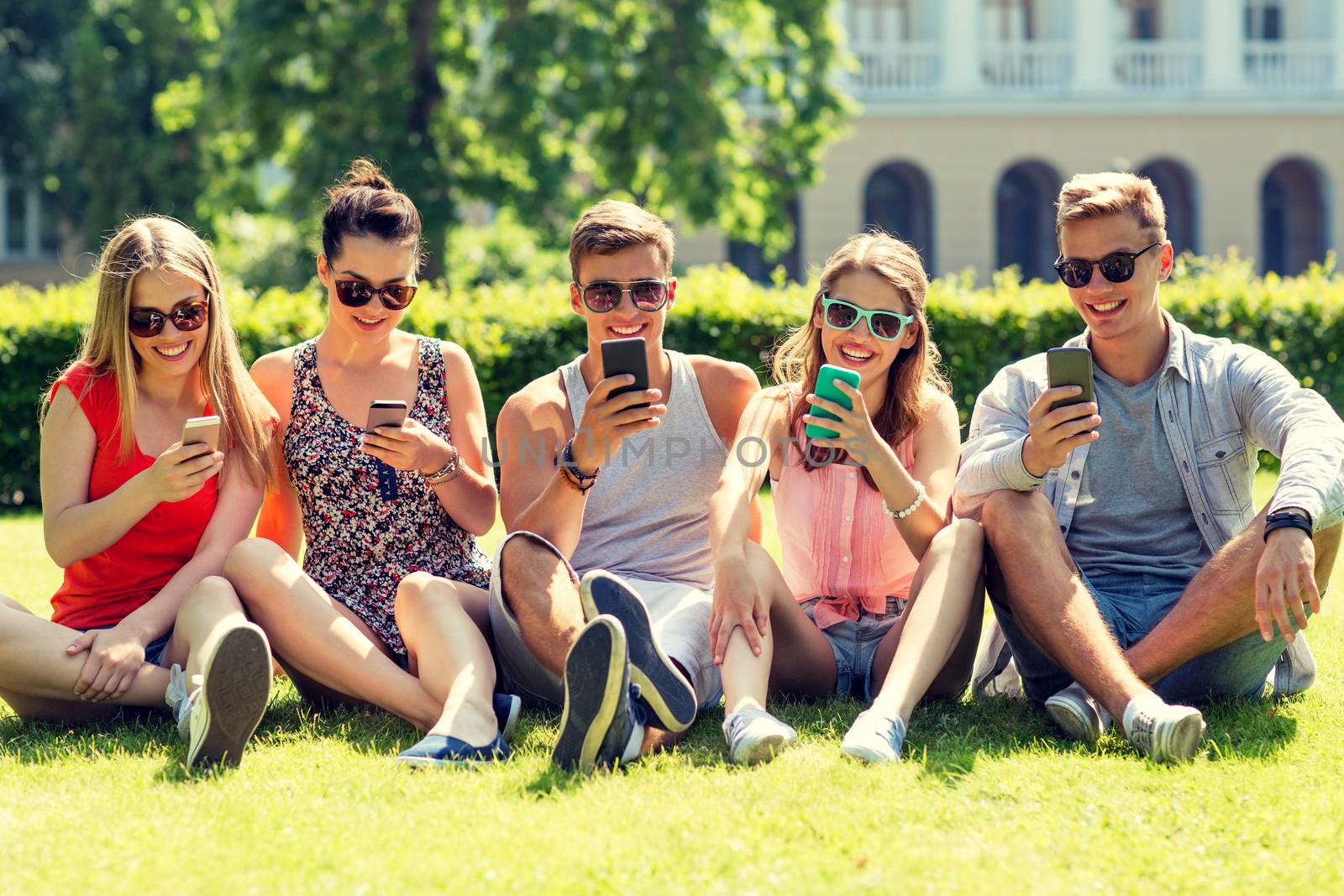 smiling friends with smartphones sitting on grass by dolgachov
