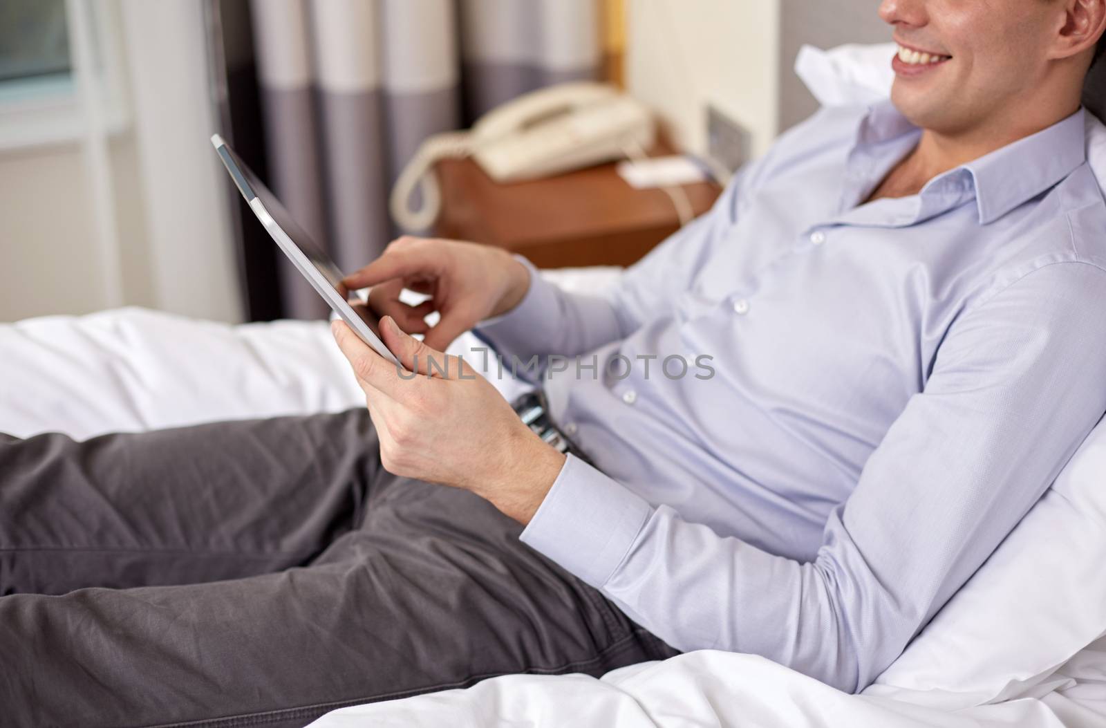 business trip, technology, internet and people concept - smiling businessman with tablet pc computer lying on bed at hotel room