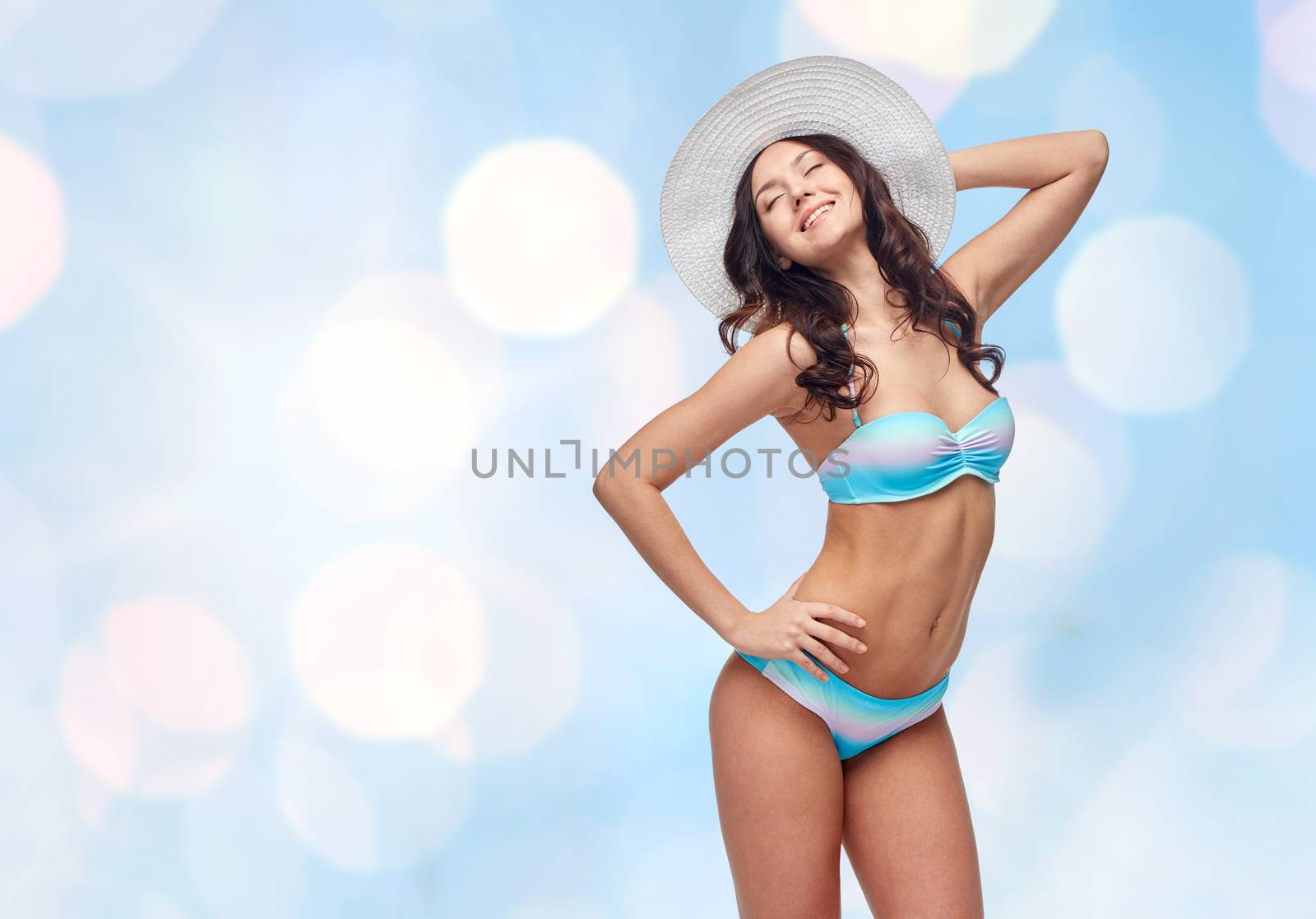 people, fashion, swimwear, summer and beach concept - happy young woman in bikini swimsuit and sun hat over blue holidays lights background