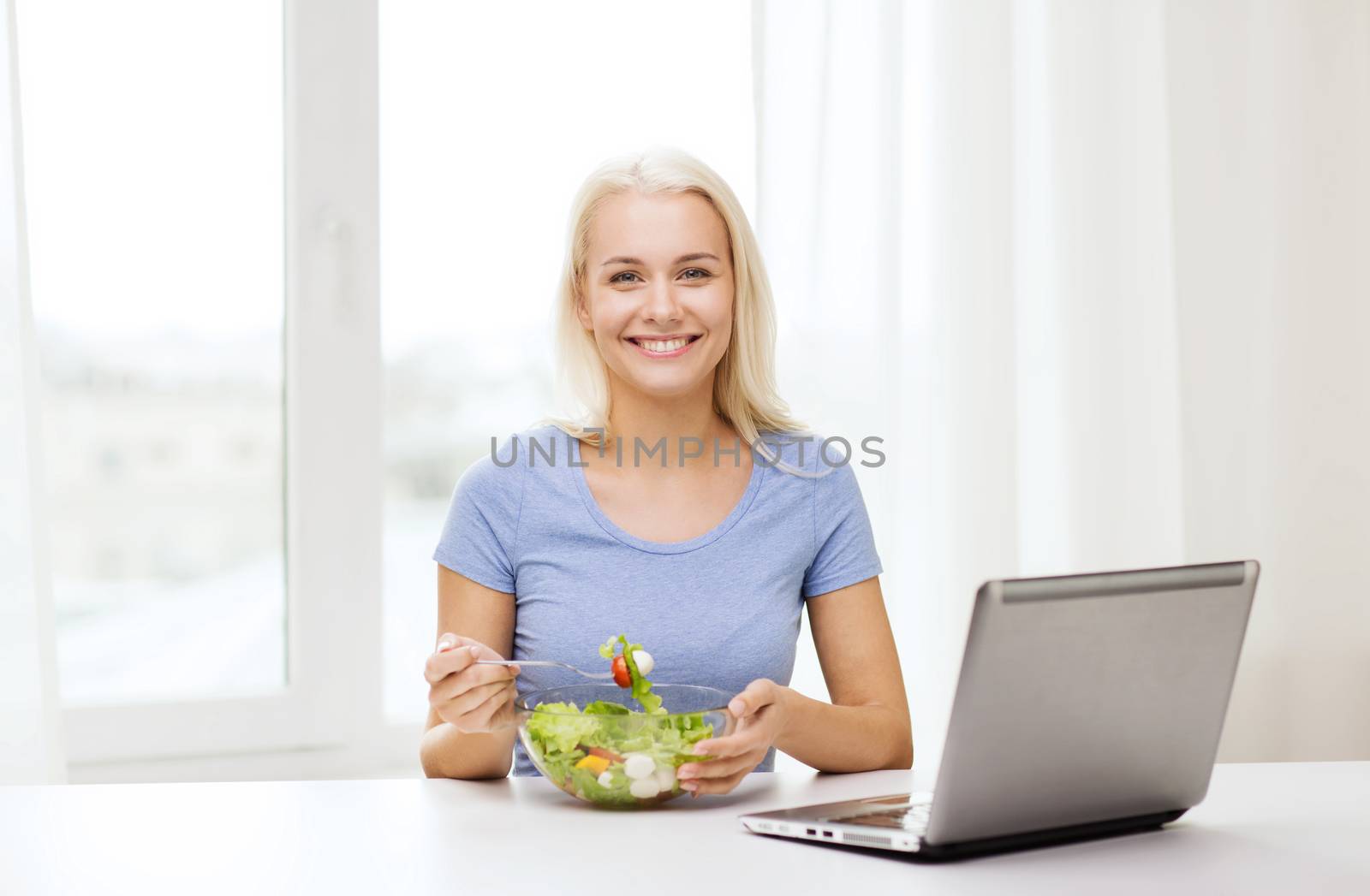 smiling woman eating salad with laptop at home by dolgachov