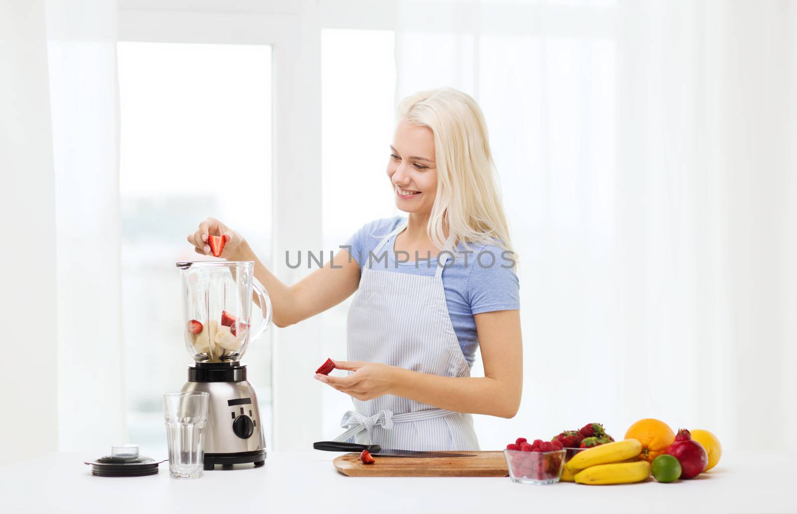 healthy eating, cooking, vegetarian food, dieting and people concept - smiling young woman putting fruits and berries for fruit shake to blender shaker at home