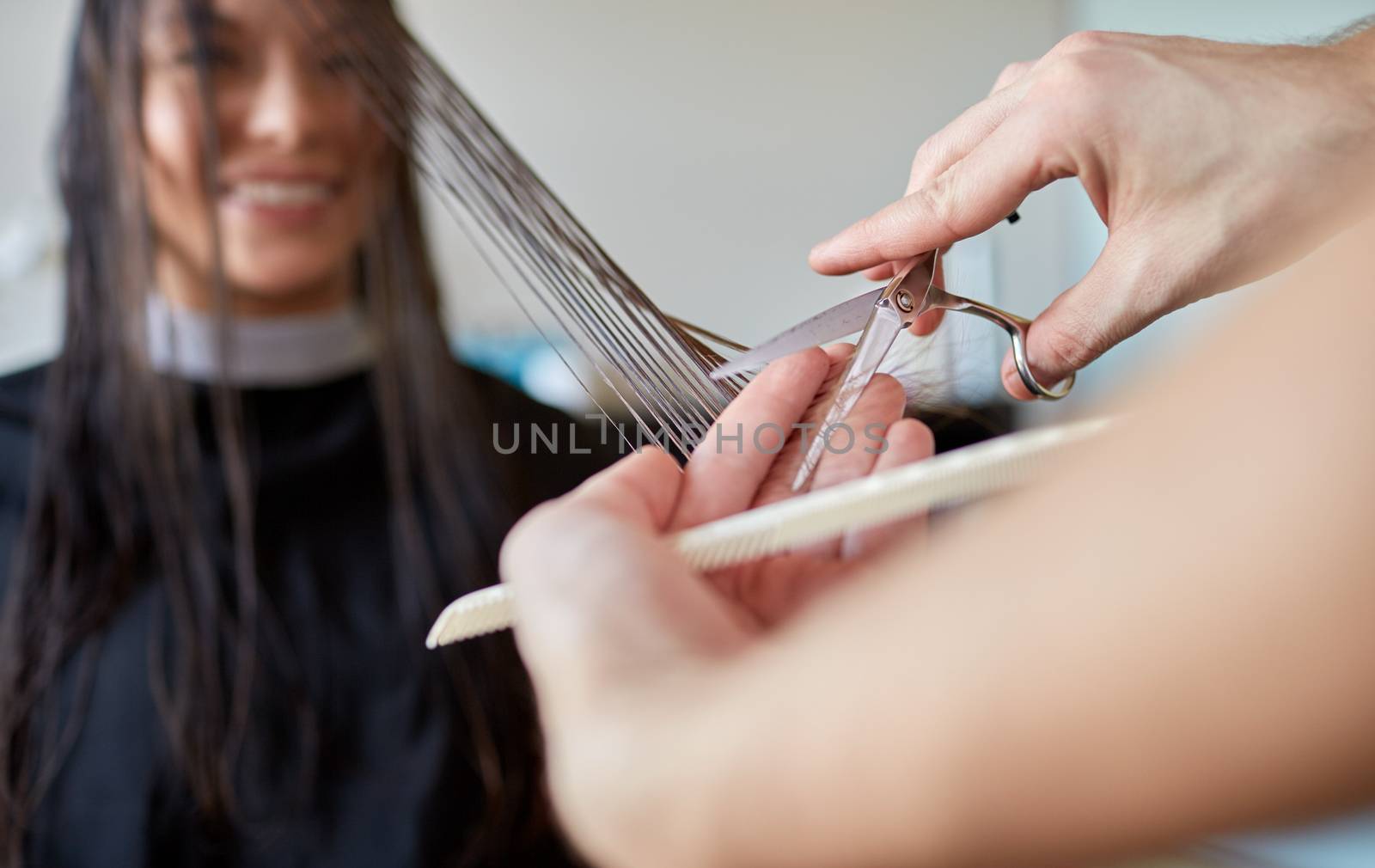 beauty, hairstyle and people concept - happy young woman and hairdresser hands with scissors and comb cutting hair tips at salon