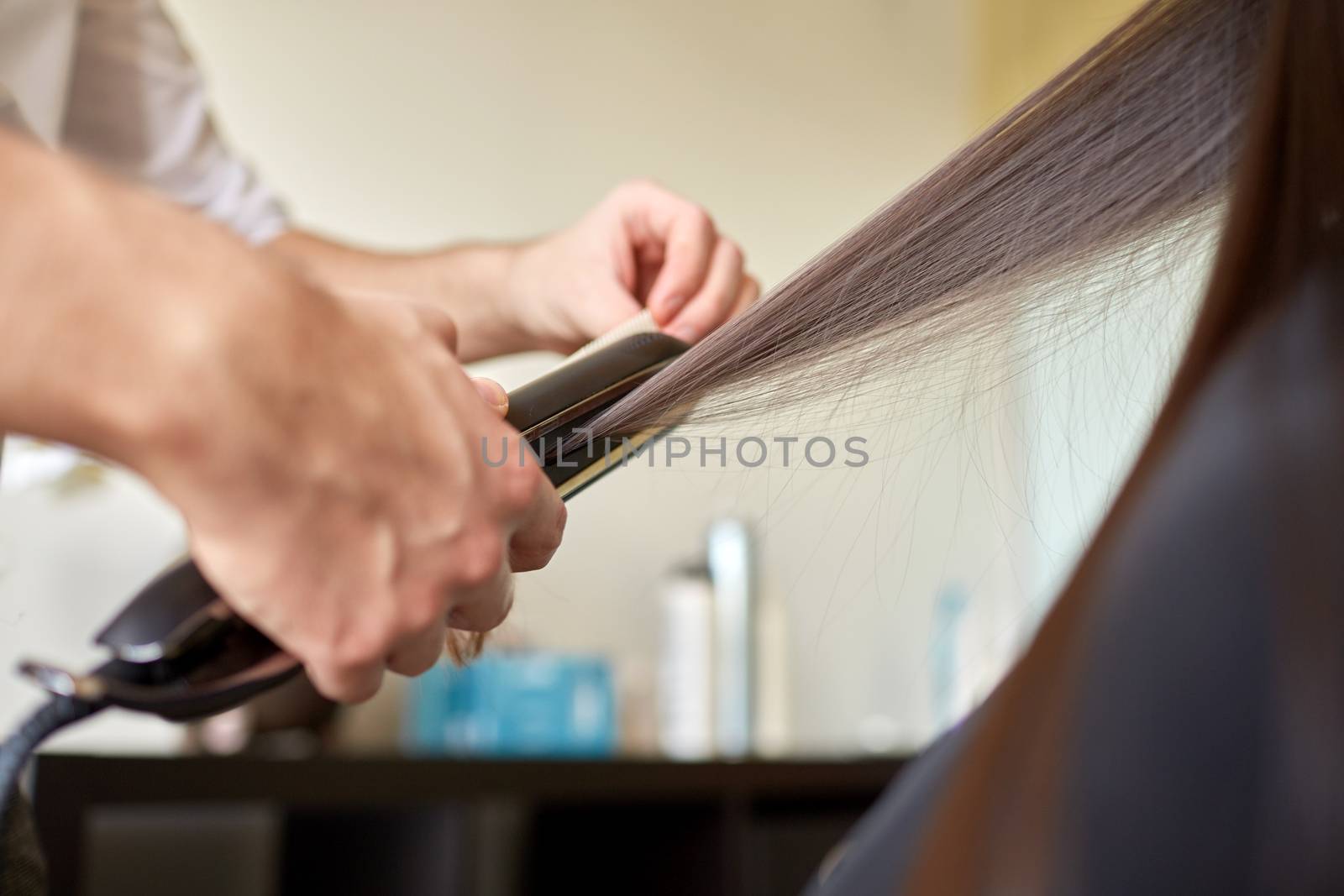 beauty, hairstyle, hot styling and people concept - close up of stylist hands with styling iron straightening woman hair at salon