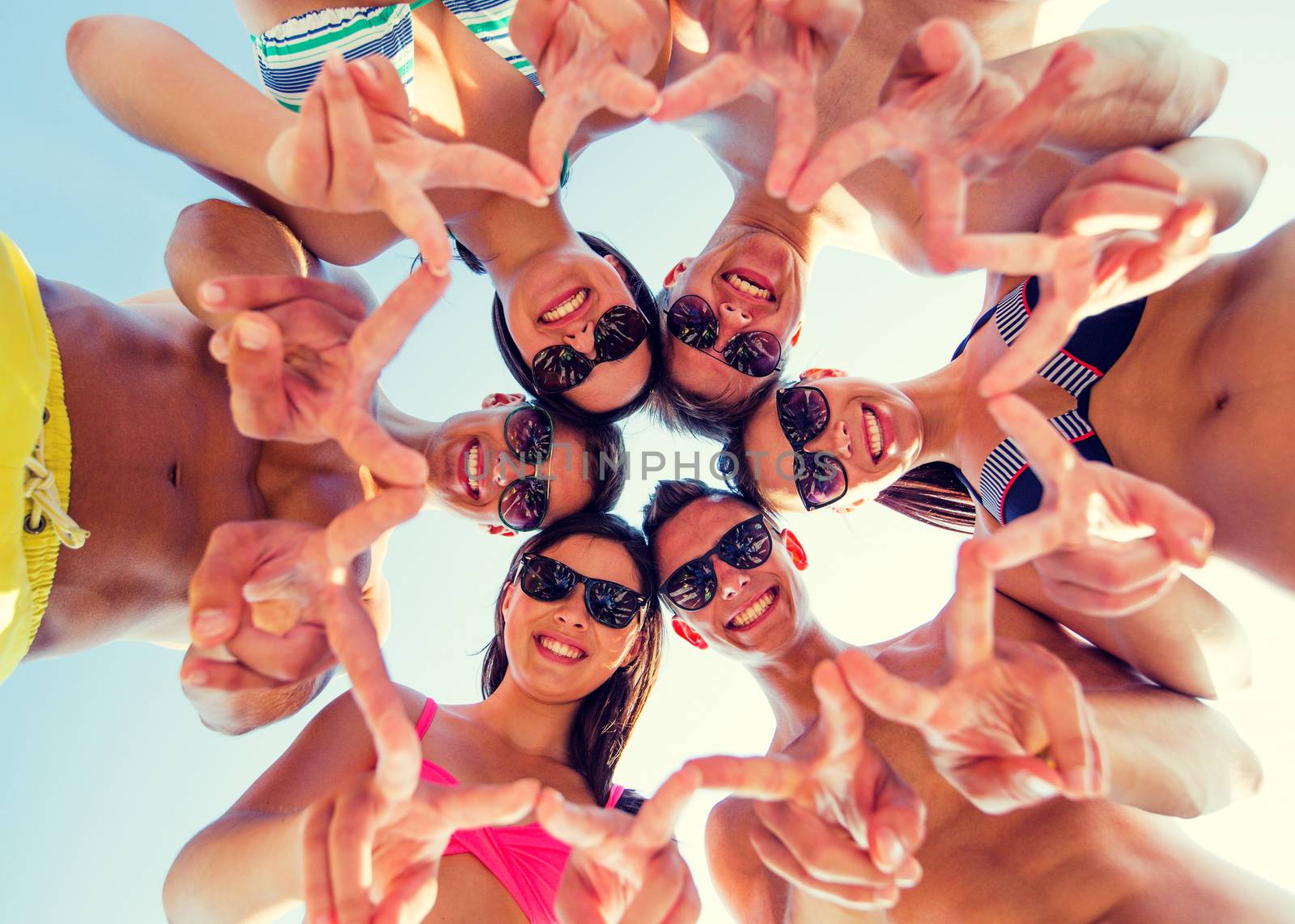 friendship, happiness, summer vacation, holidays and people concept - group of smiling friends wearing swimwear and showing victory or peace sign standing in circle over blue sky