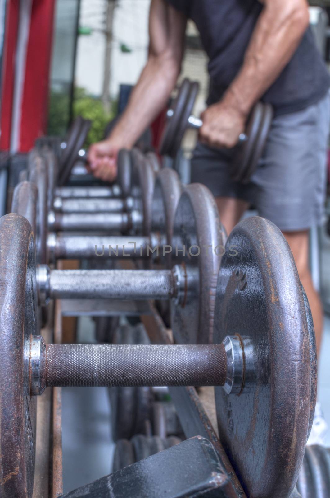 Focus on rusty dumbbell weight on rack in foreground while unrecognized man in background picks up dumbbells in gym