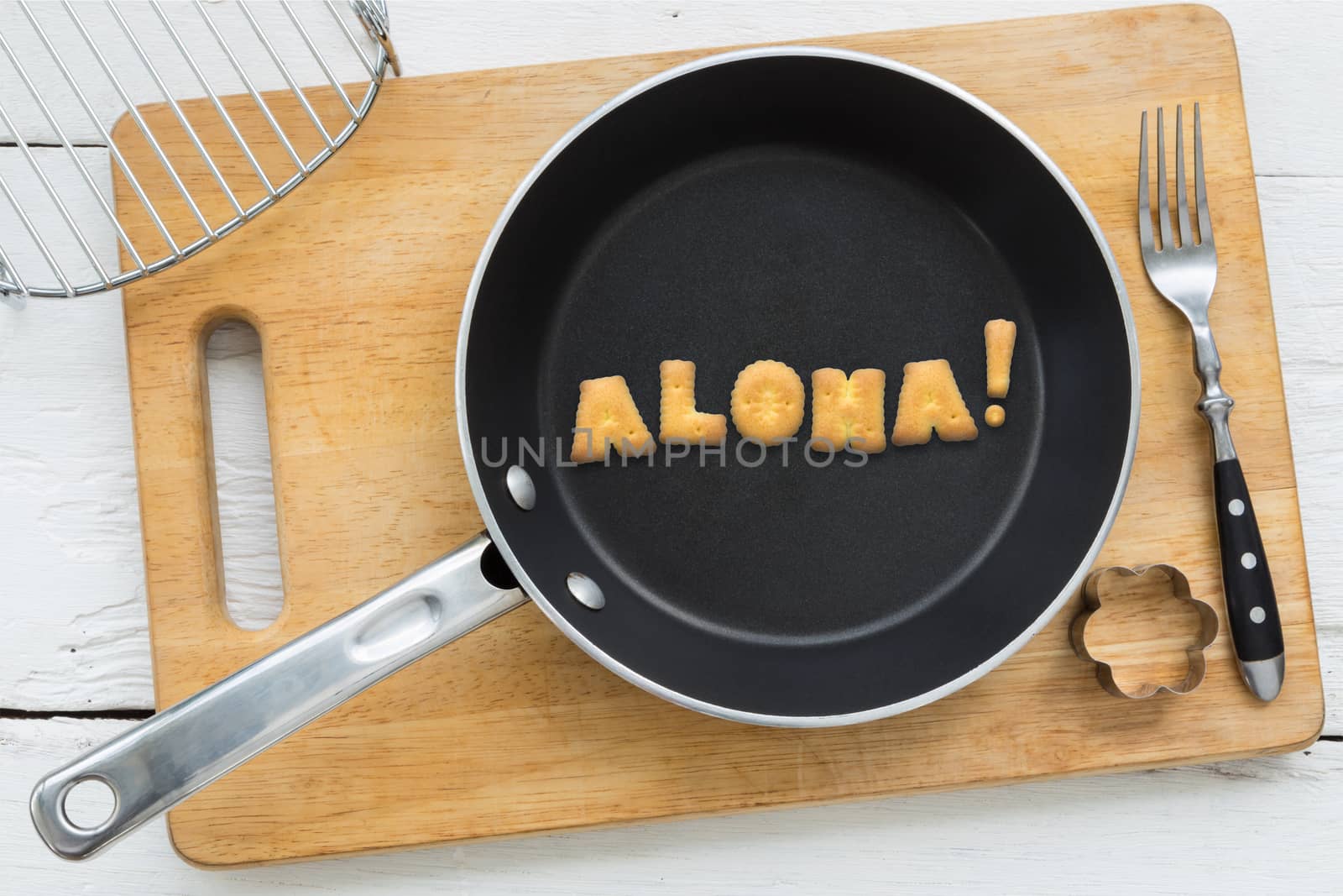 Top view of alphabet collage made of biscuits. Word ALOHA putting in black pan. Other kitchenware: fork, cookie cutter and chopping board putting on white wooden table, vintage style image.