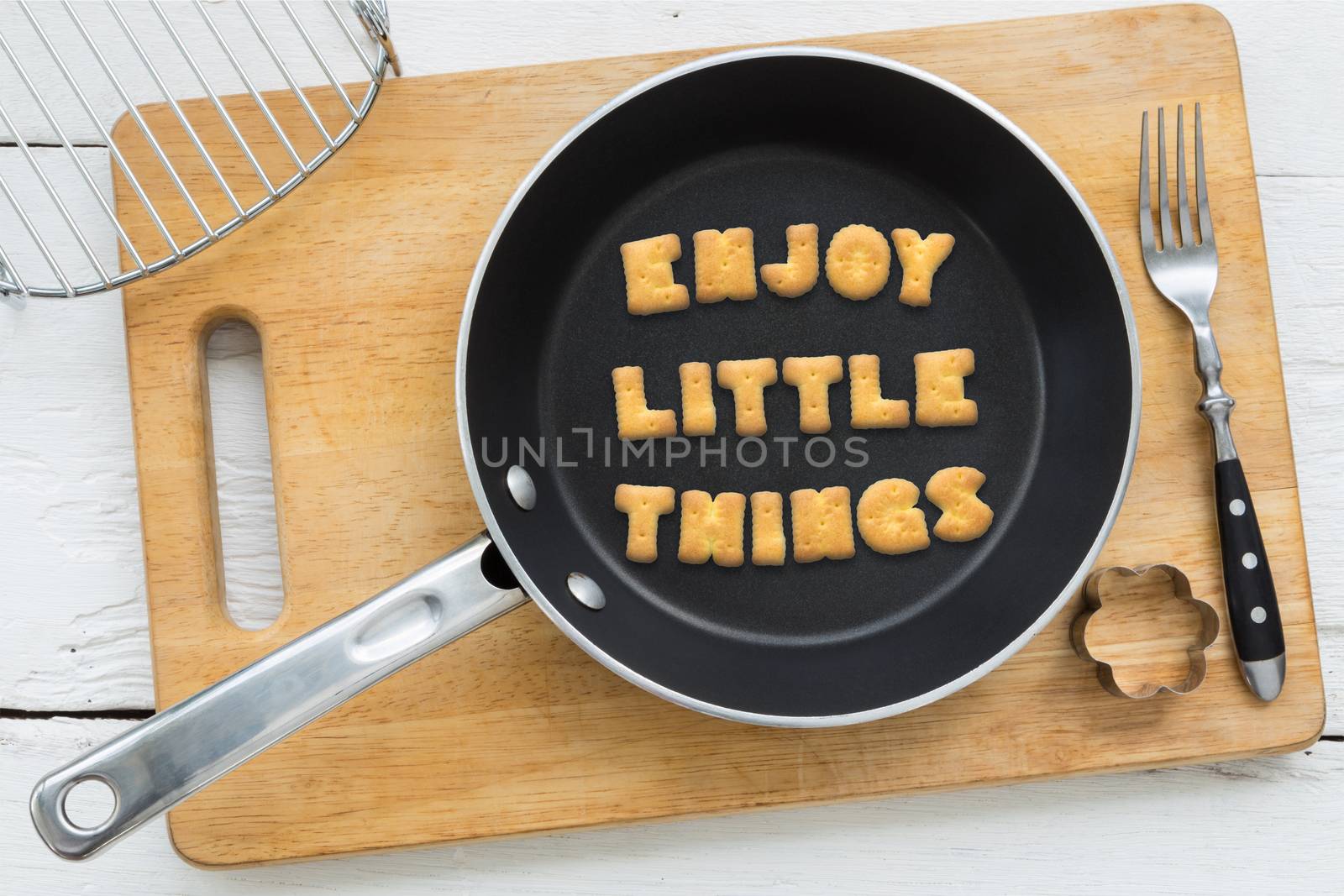 Top view of alphabet collage made of crackers. Quote ENJOY LITTLE THINGS putting in black pan. Other kitchenware: fork, cookie cutter and chopping board putting on white wooden table, vintage style image.