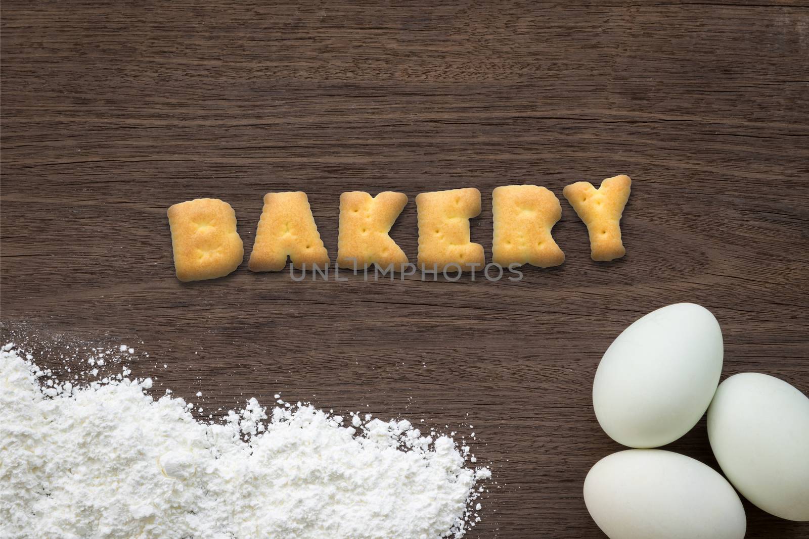 Top view of letter text collage made of cookies biscuits. Word BAKERY putting on baking background with eggs and flour on vintage natural wood kitchen table.