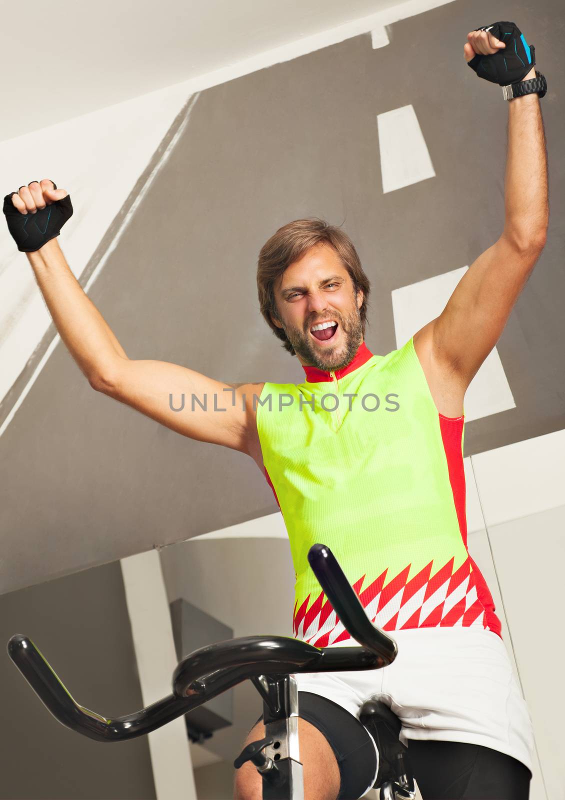 Young happy sportive man in a jim spinning a cycle, arms up smiling excited. Dressed in a sportive wear.