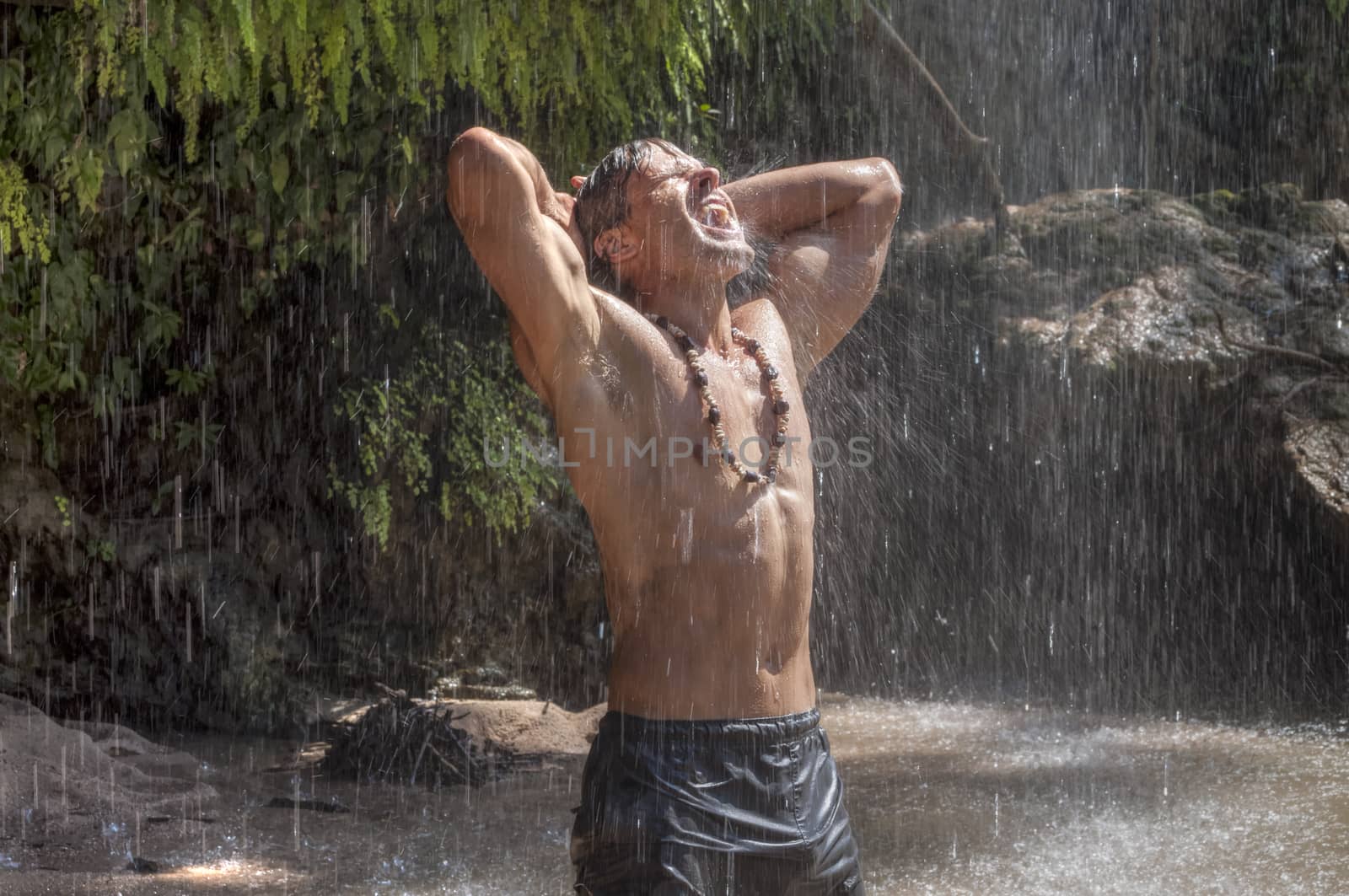 Handsome muscular shirtless Caucasian man stretches arms behind head and opens mouth to catch raindrops from waterfall in tropical paradise in Chiapas, Mexico