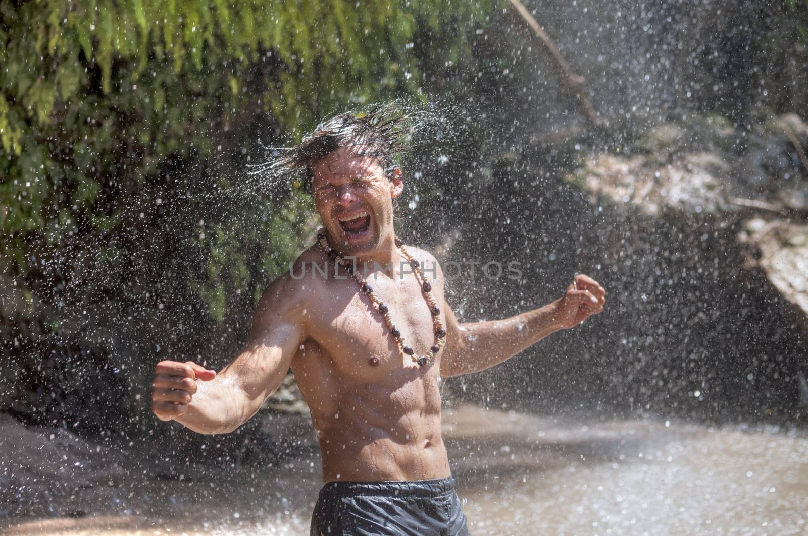 Handsome muscular shirtless Caucasian man flexes and shakes water from hair in euphoric excitement as he stands under raining tropical waterfall in river
