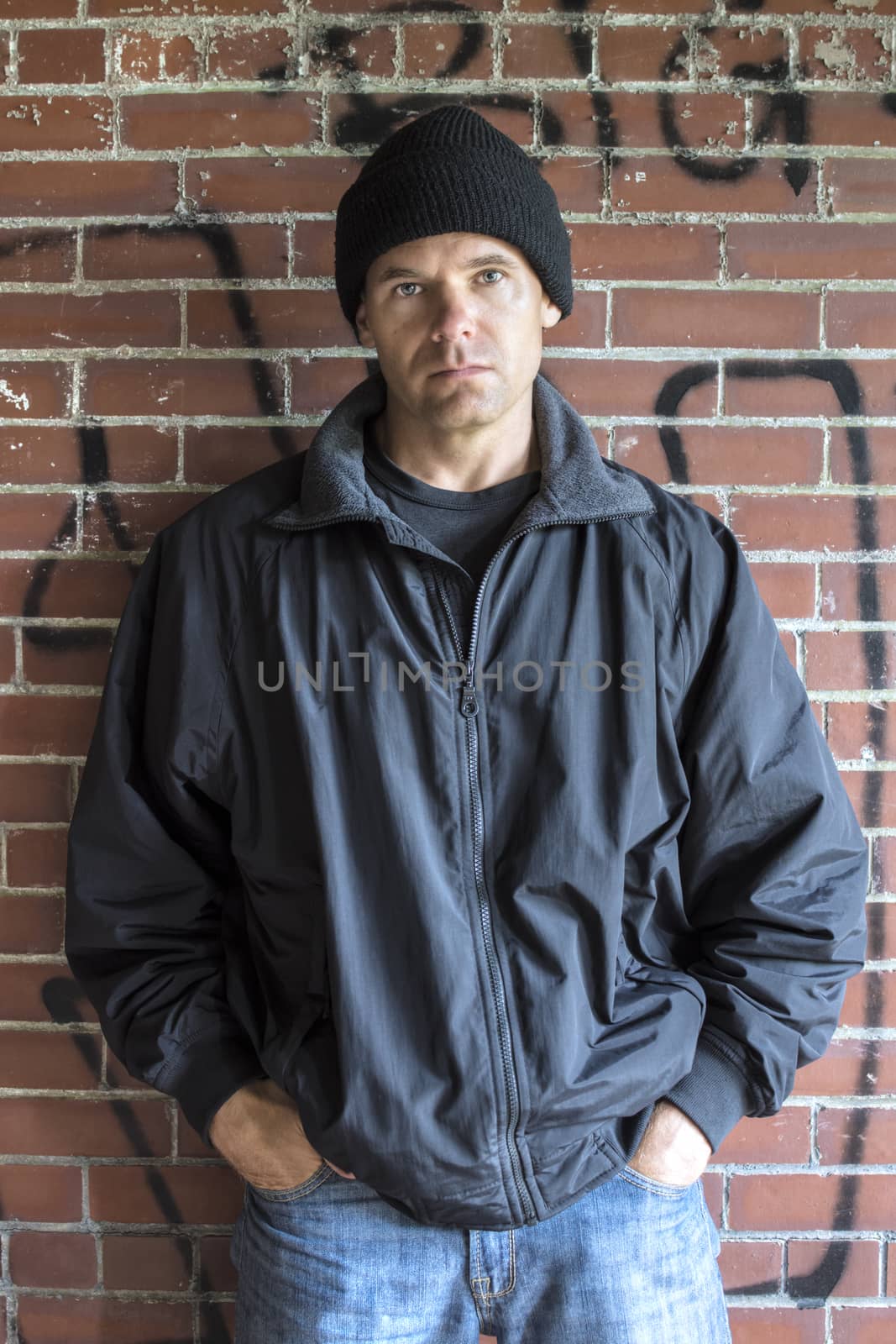 Caucasian man with serious expression wears hoodie, jacket and jeans and leans against brick wall with graffiti in urban setting with hands in pockets