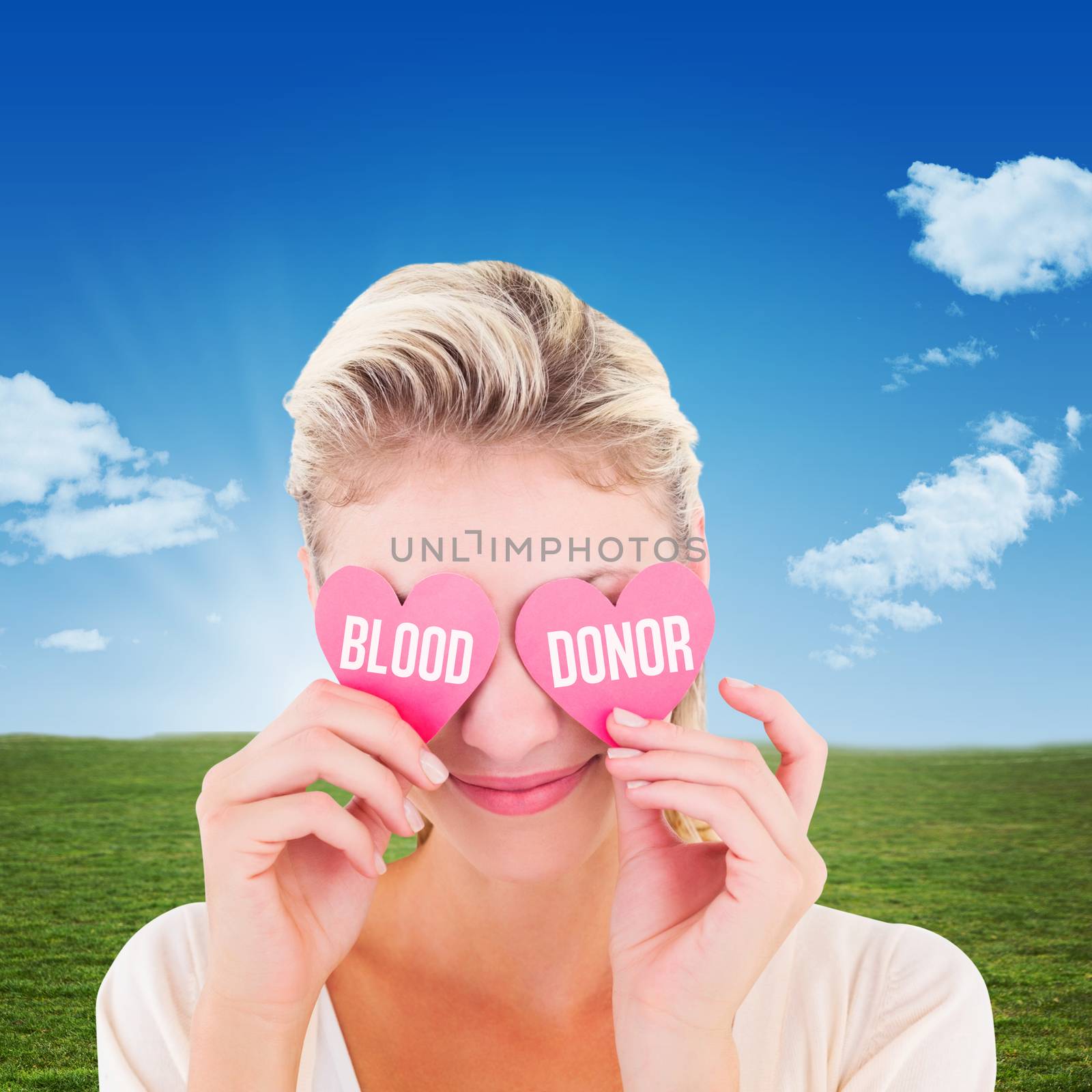 Attractive young blonde holding hearts over eyes against blue sky over green field
