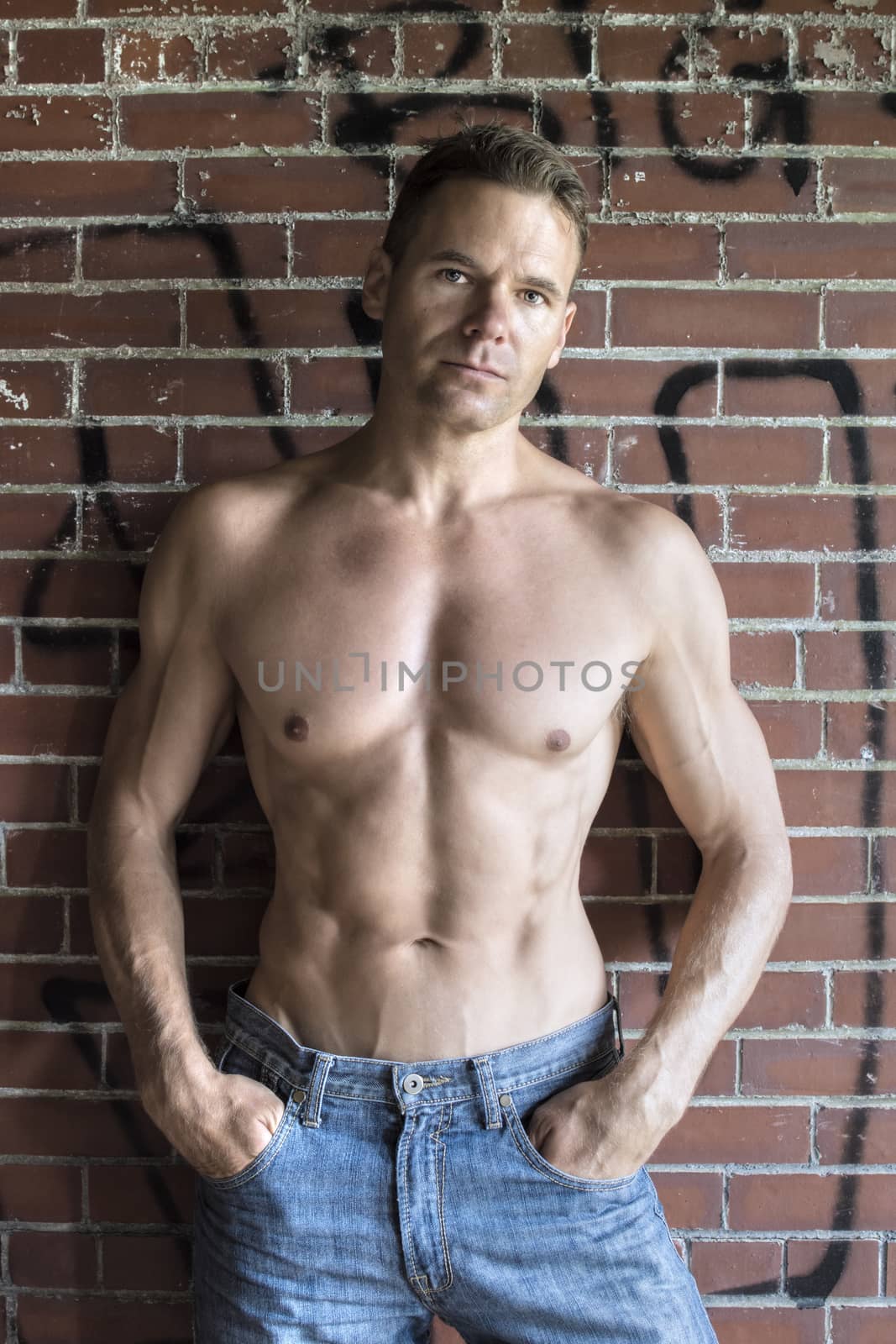 Muscular shirtless Caucasian man in blue jeans standing against brick wall with graffiti in urban setting