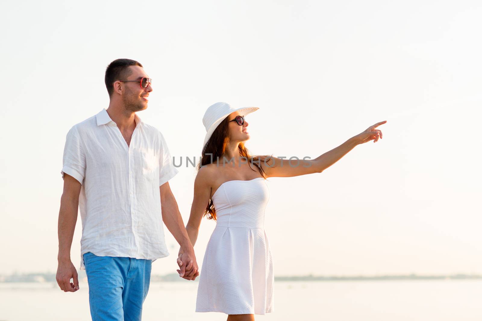 love, travel, tourism, gesture and people concept - smiling couple on summer vacation wearing sunglasses and pointing finger walking at seaside