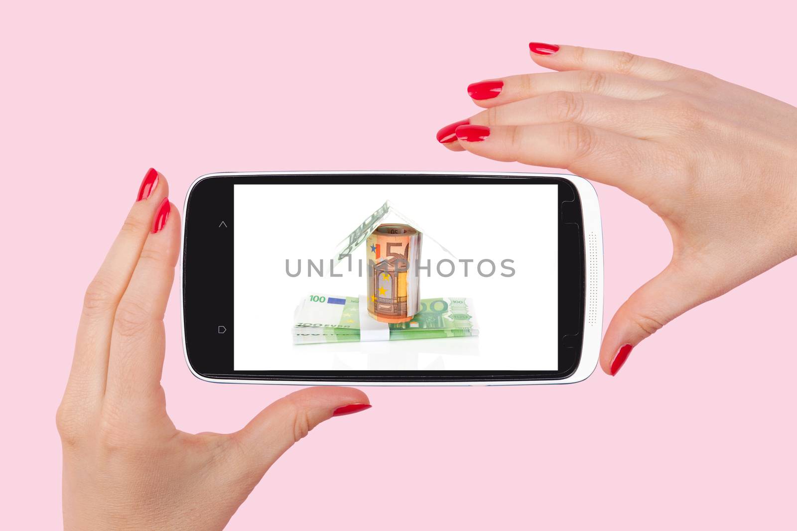 Mortgage at your fingertips. Female hand holding a smartphone with house made from bills isolated on pink background.
