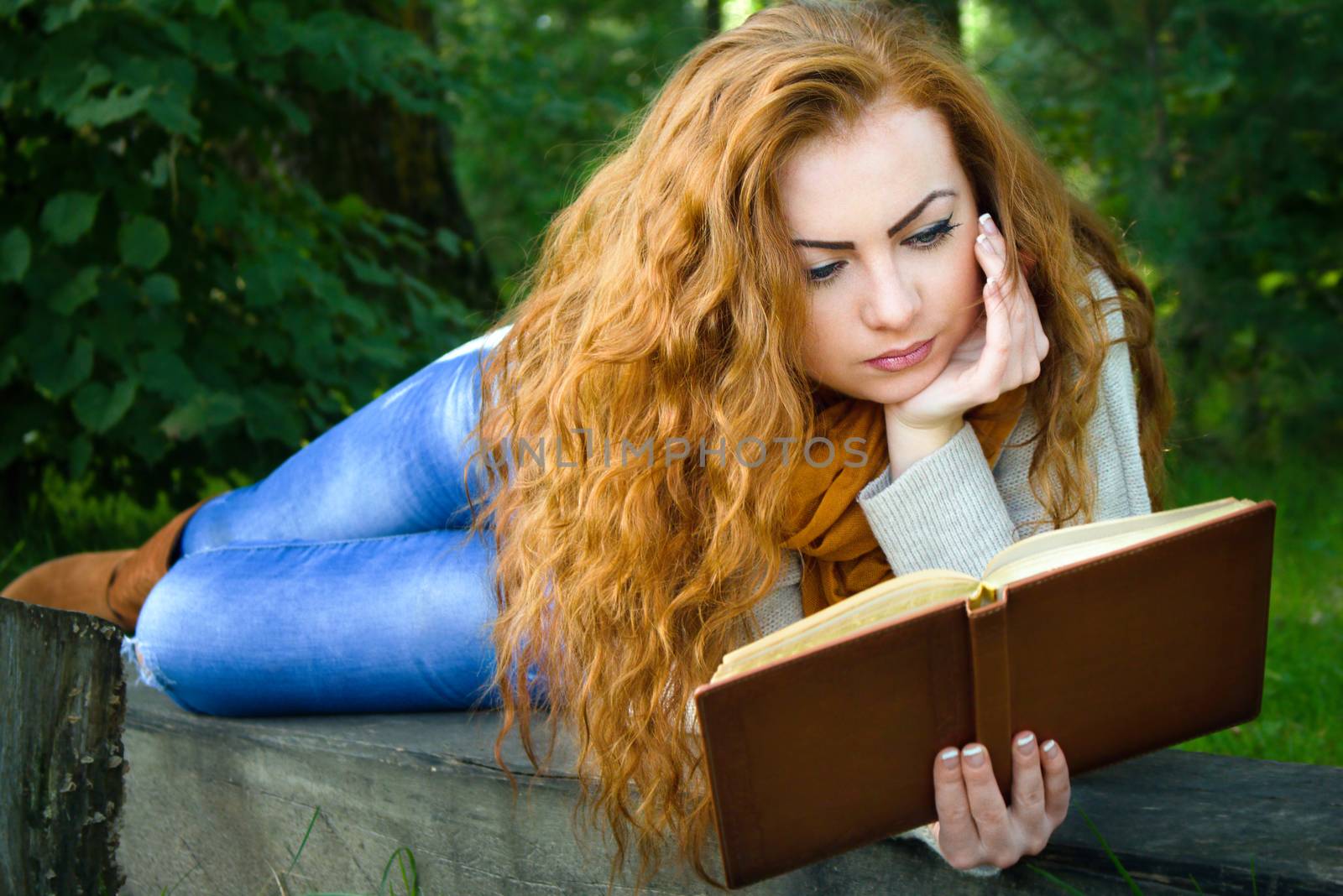 Beautiful ginger-haired woman reading a book in park lying on the bench