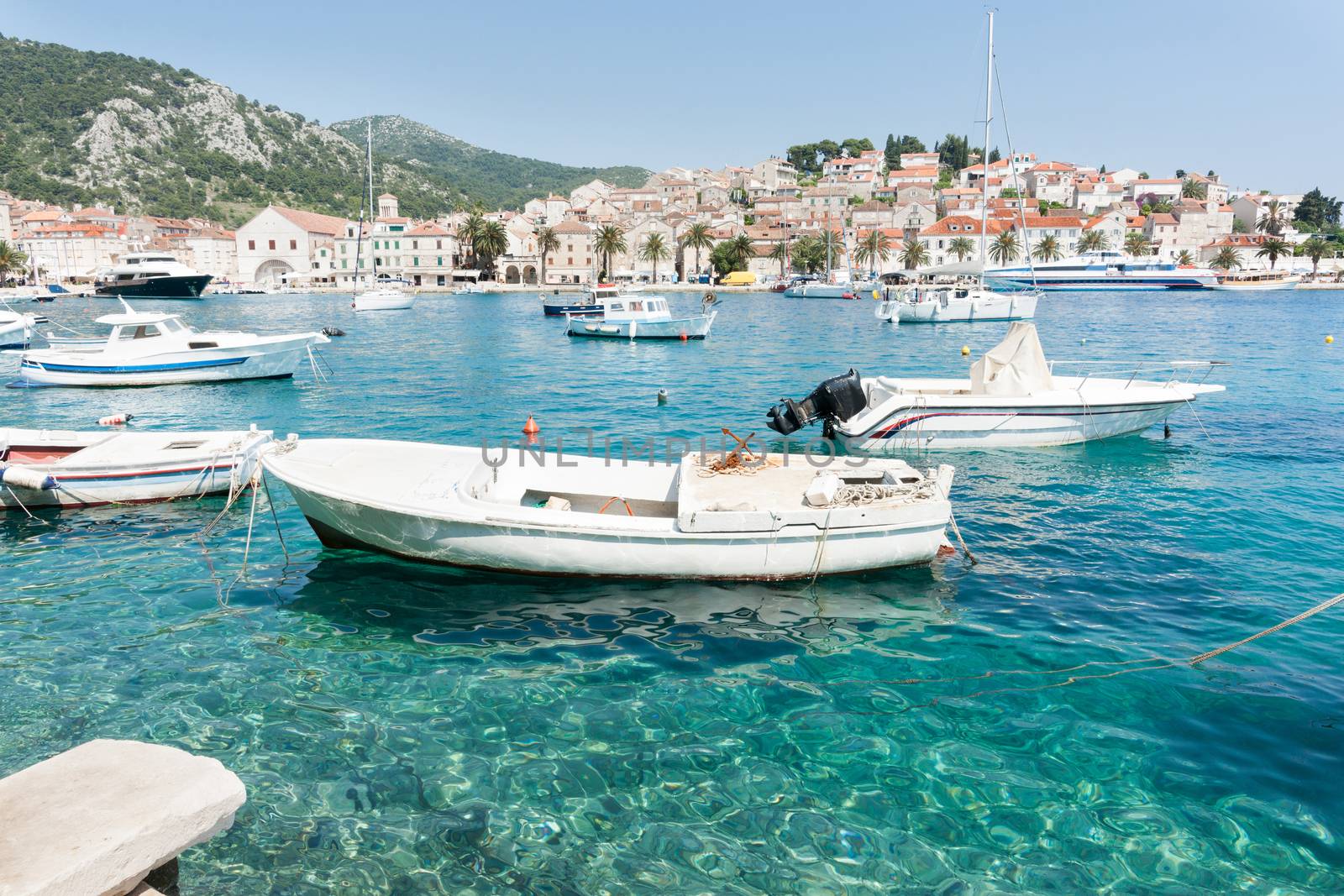 Boats in the harbour with historic town with its build white buildings with terracotta tiles around the shore and rolling green hills.,Hvar, Croatia.