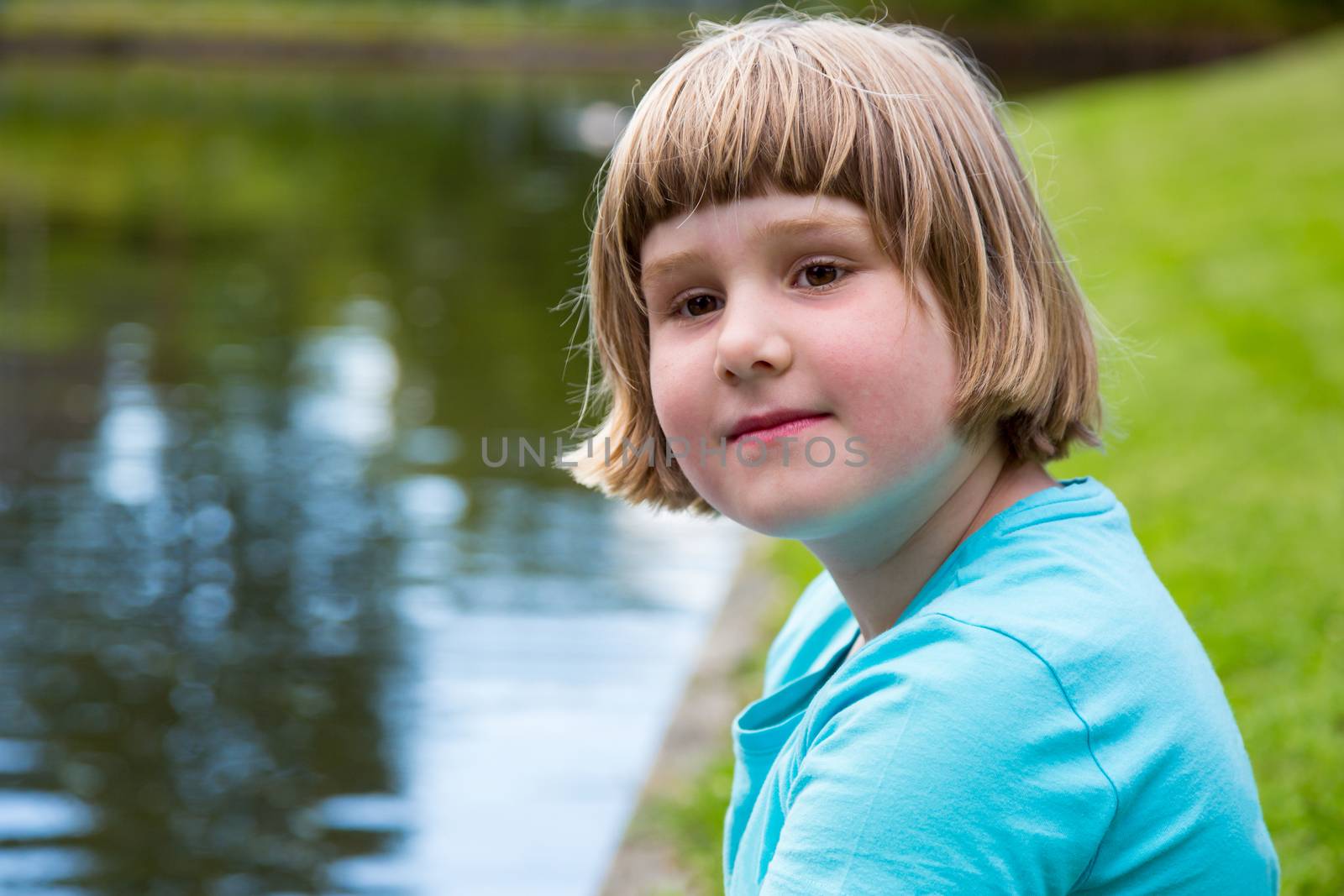 Young blonde dutch girl sitting at water of pond in park