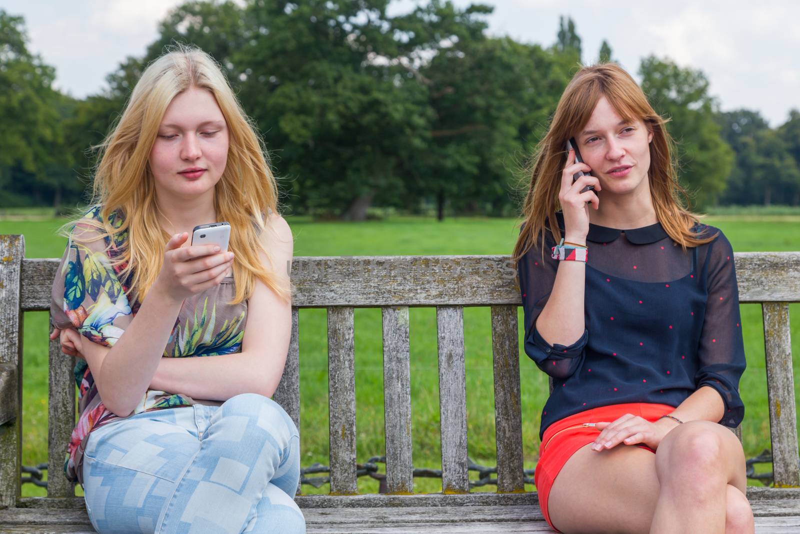 Two caucasian teenage girlfriends sitting on wooden bench in park calling with mobile phone