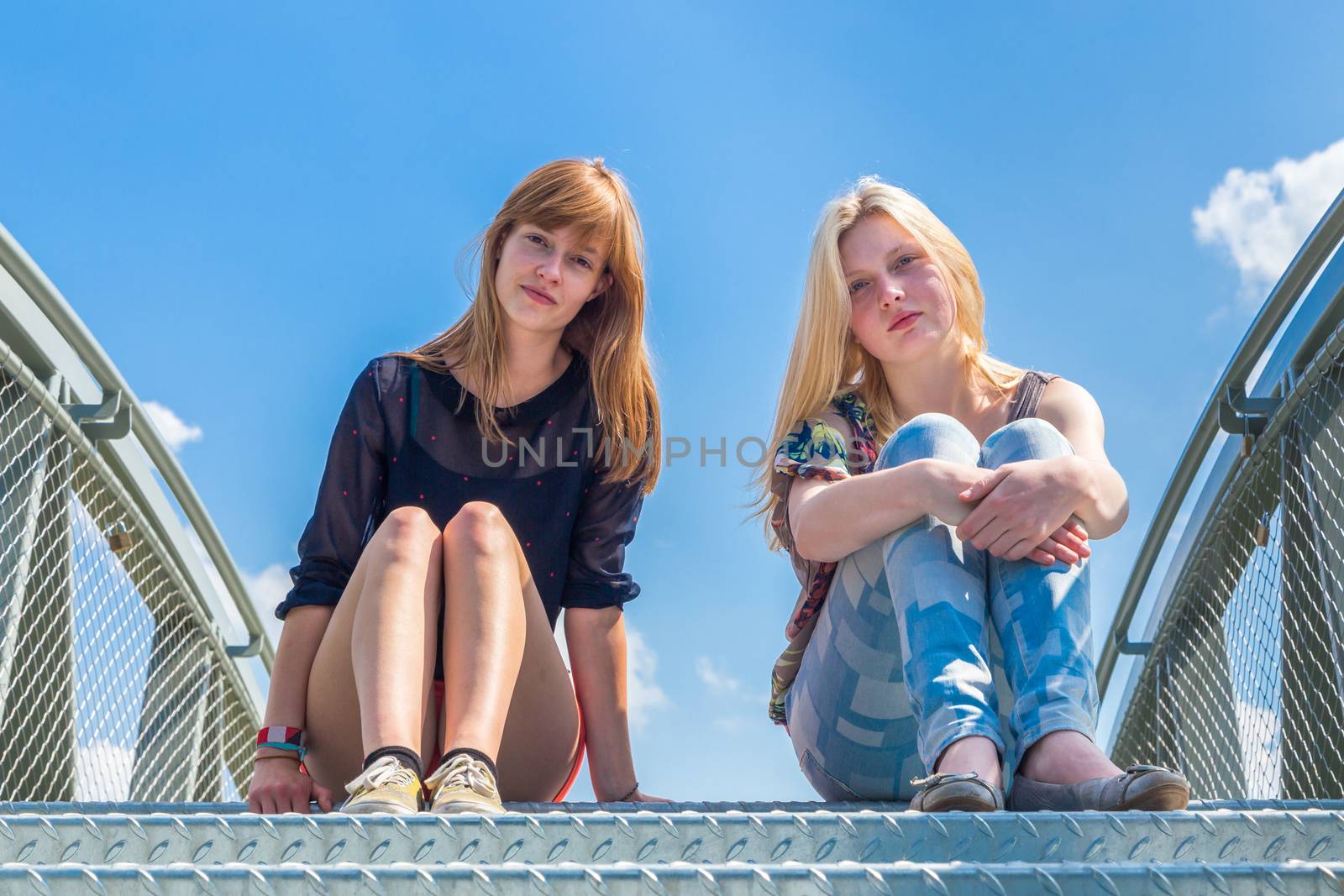 Two girls sitting on metal bridge with blue sky by BenSchonewille