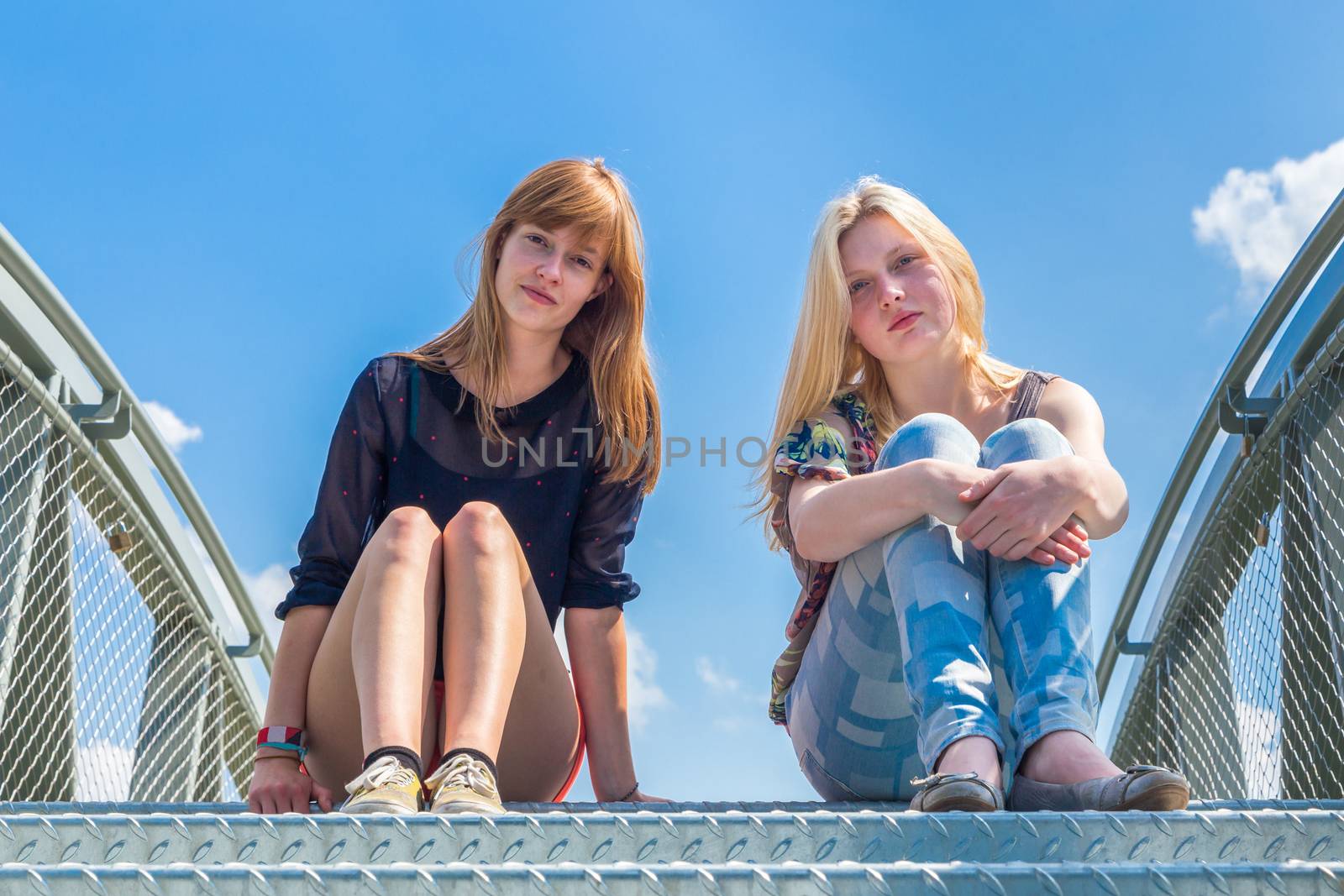 Two dutch teenage girlfriends sitting on metal bridge with blue sky