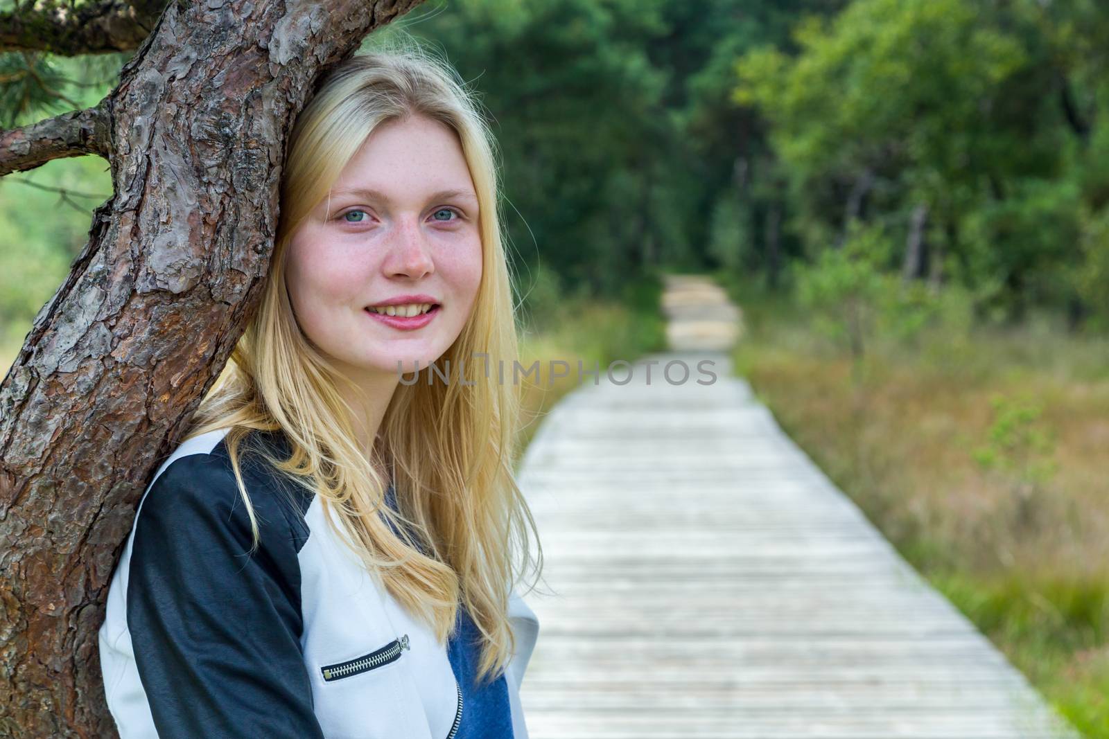 Portrait of blonde teenage girl with trunk and wooden footpath in forest nature