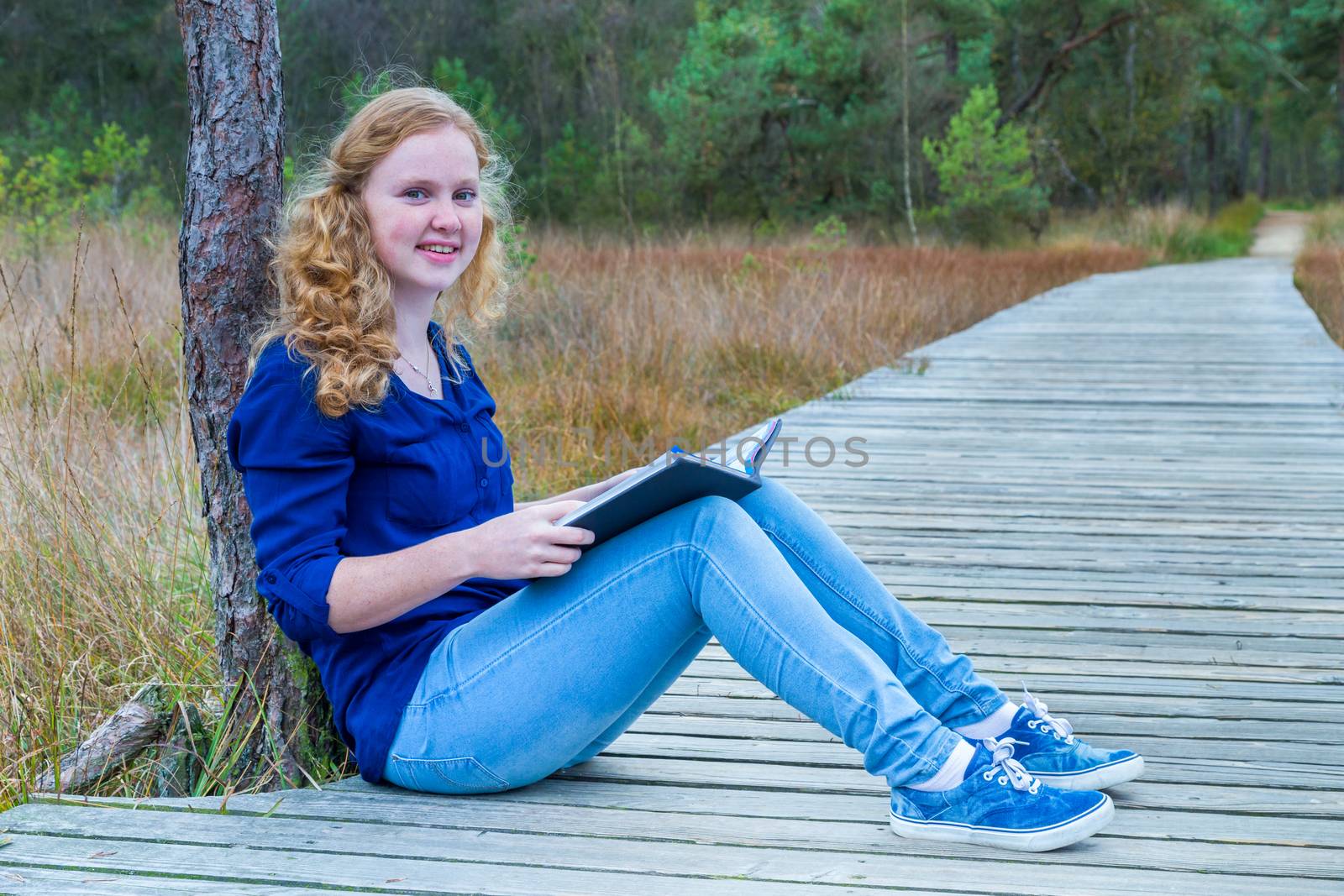 European teenage girl reading book on wooden path in forest
