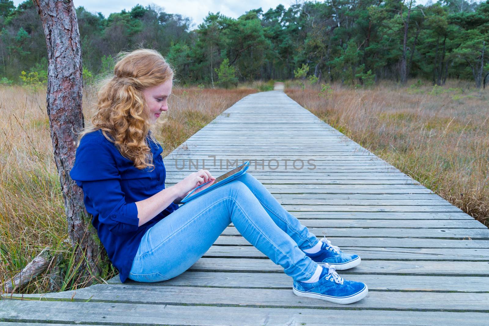Caucasian teenage girl working with tablet computer on wooden footpath in nature