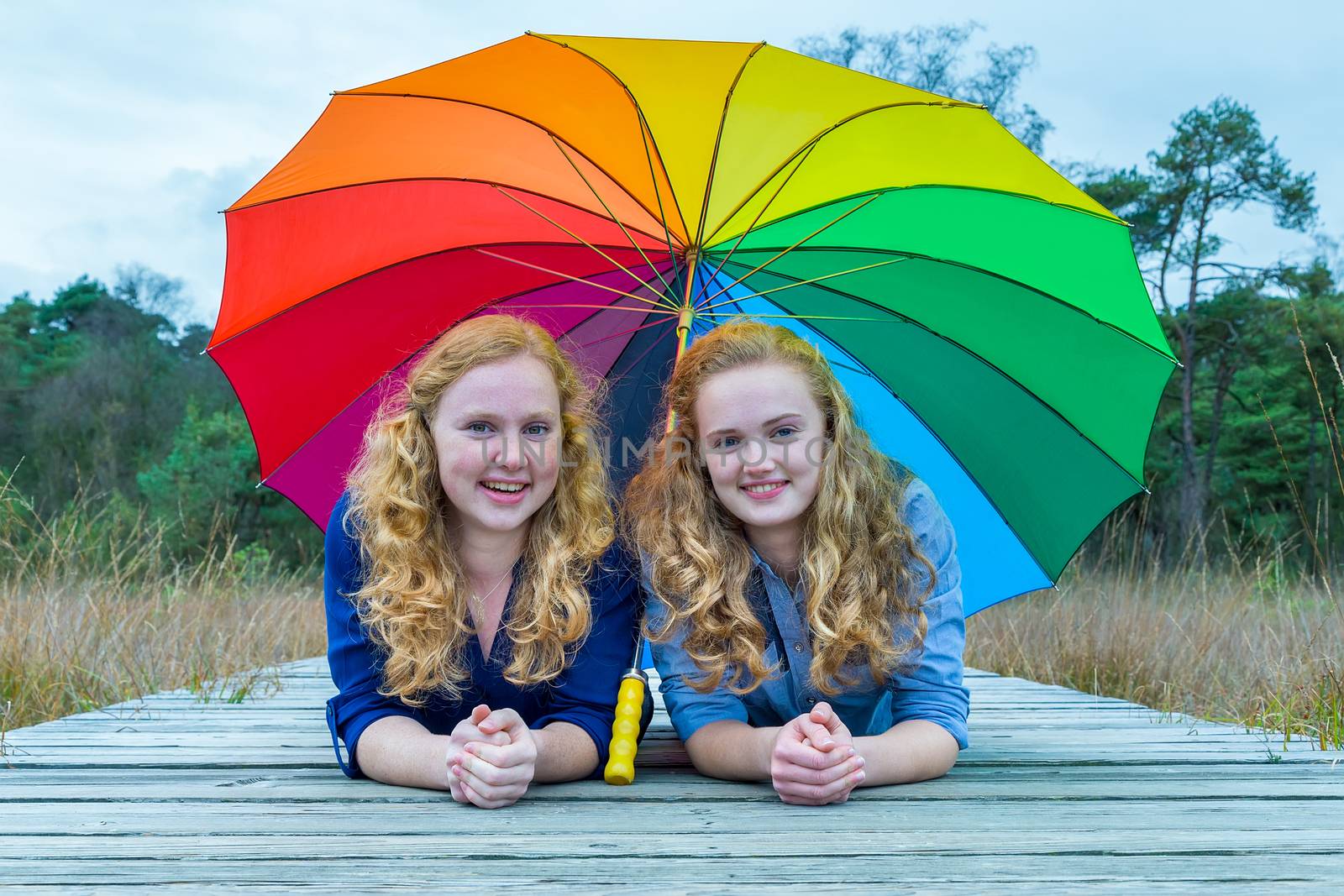 Two smiling caucasian teenage girlfriends lying in nature under colorful umbrella