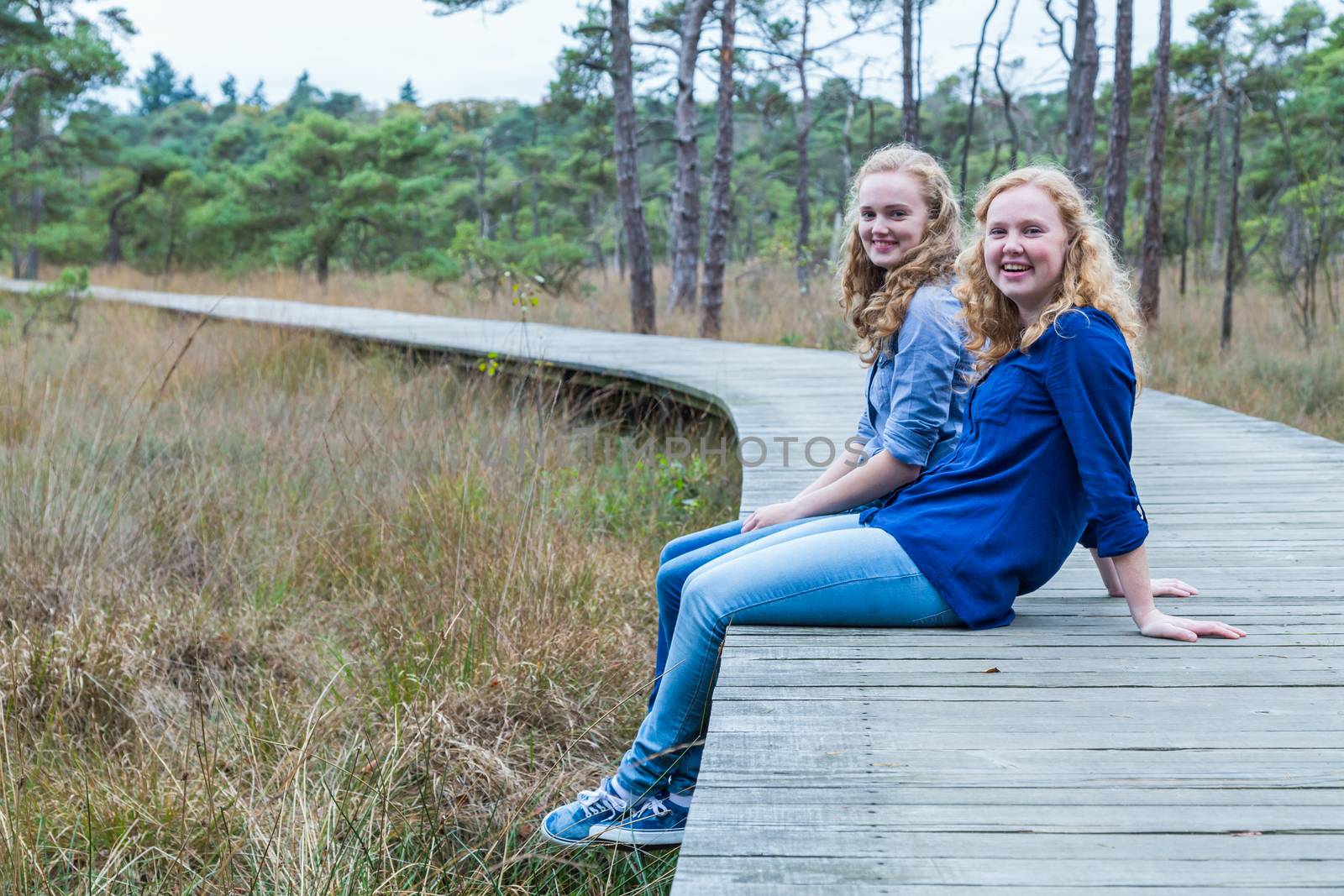 Two european teenage sisters sitting on wooden footpath in forest