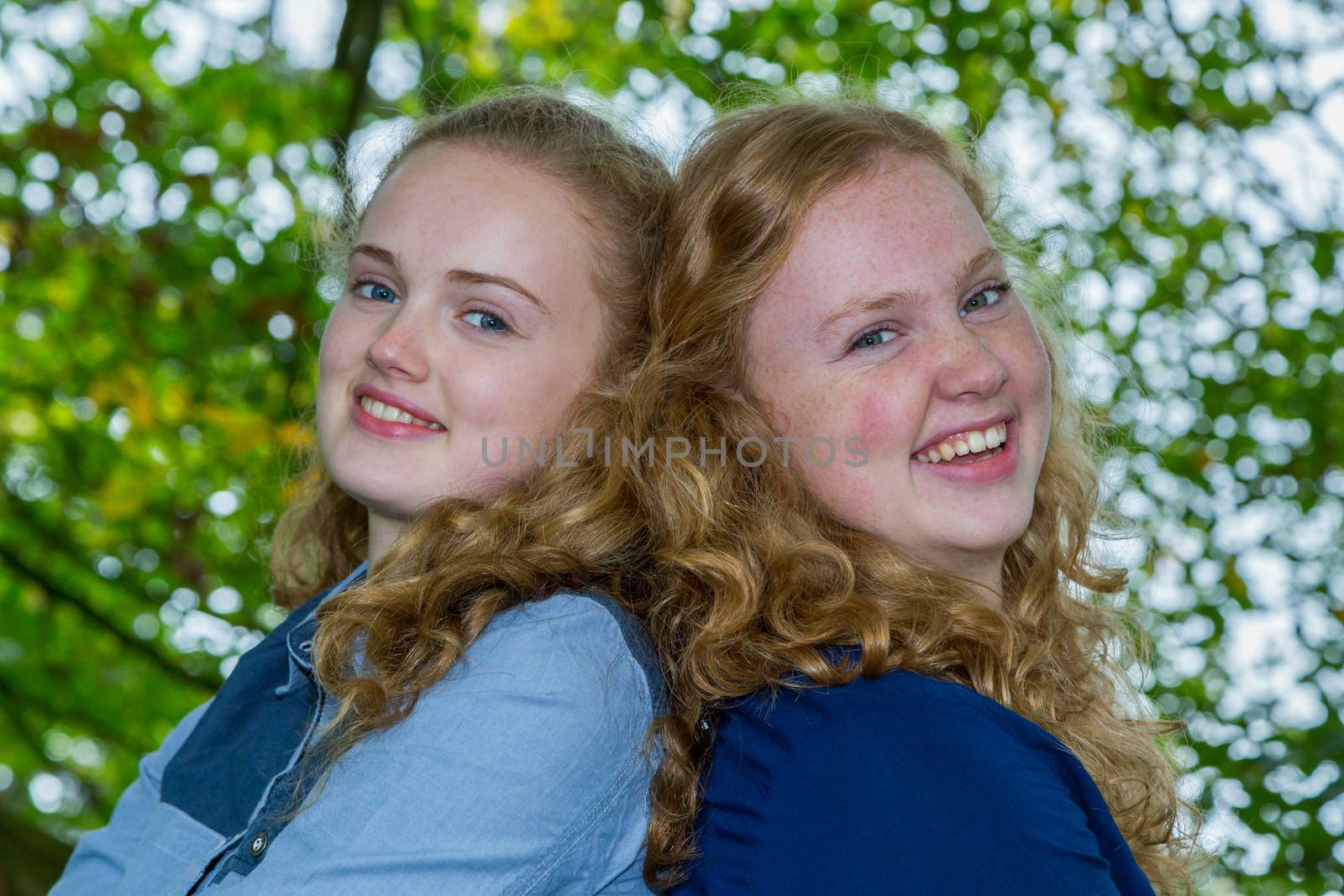 Two caucasian teenage sisters heads together laughing under green tree
