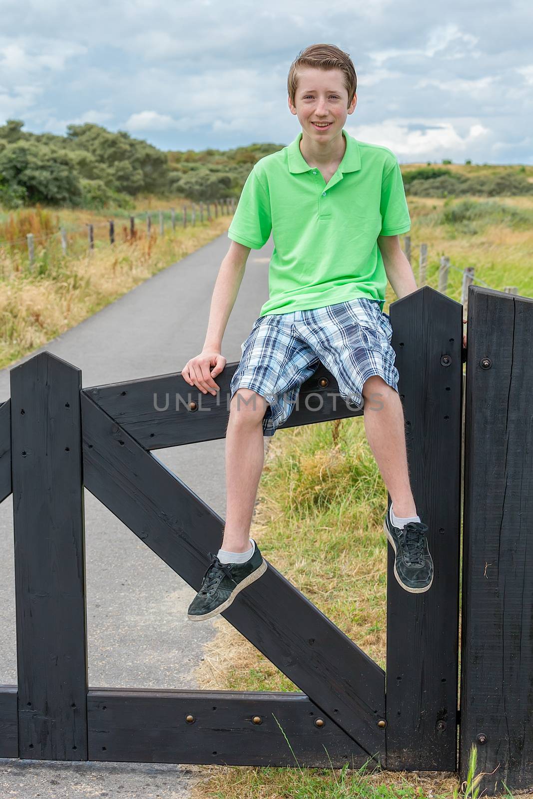 Caucasian teenage boy sitting on black wooden fence in nature as tourist enjoying vacation