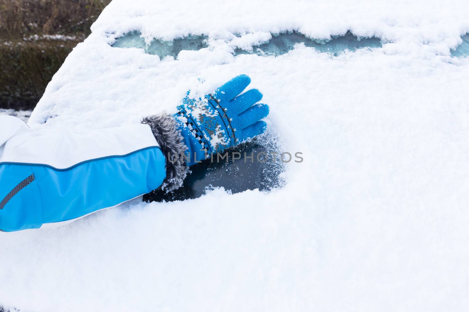 Arm with blue glove removing snow from car windowin winter season