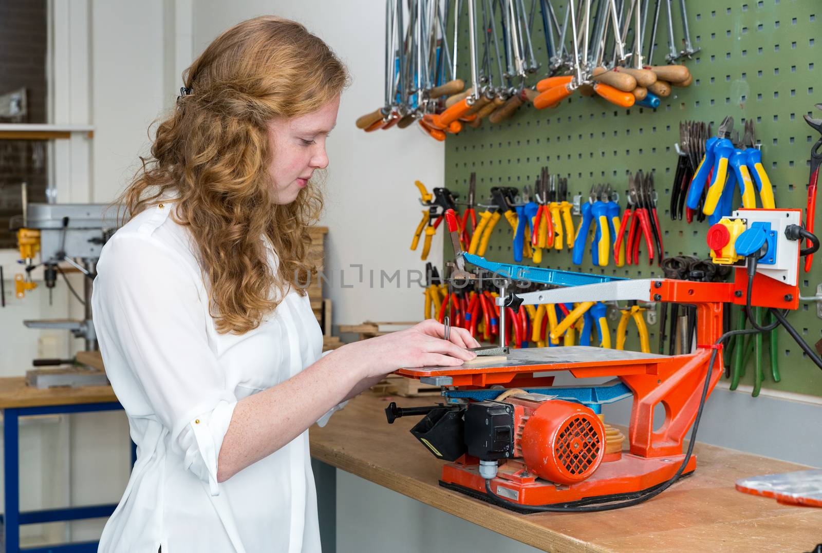 Caucasian redhead teenage girl operating electric jigsaw in classroom of school building