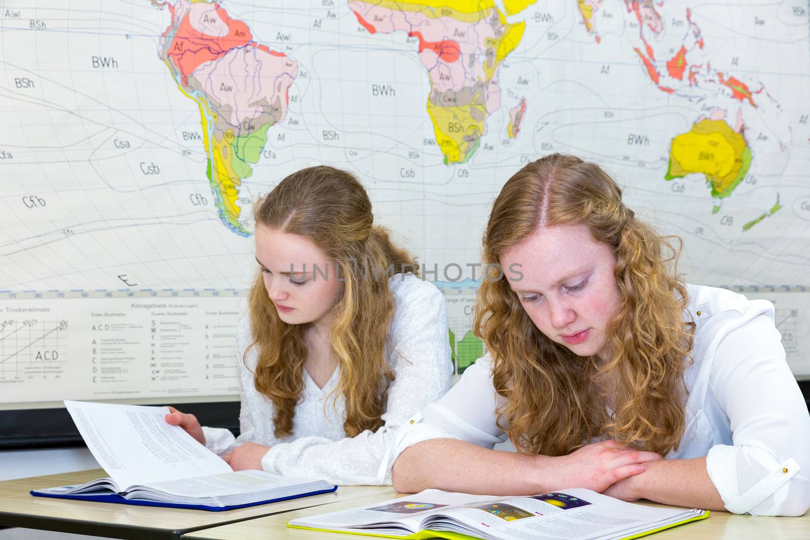 Two caucasian teenage sisters studying in front of wall world chart in classroom on school