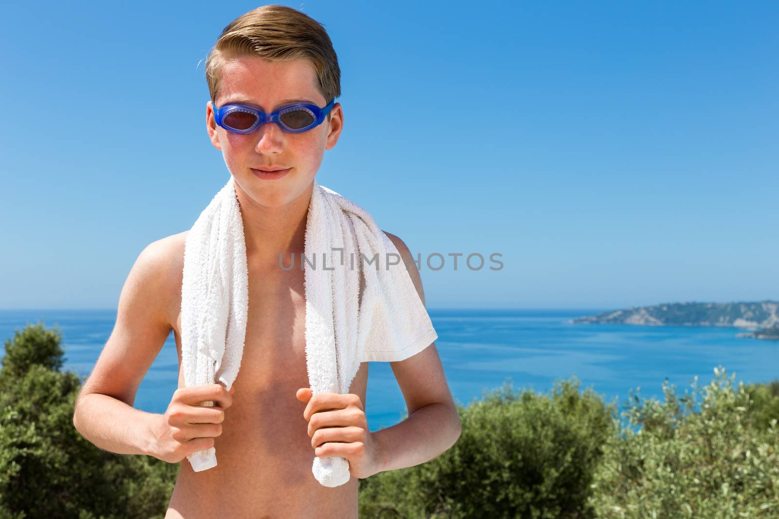 European teenage boy holding white towel  and wearing swimming goggles in front of blue sea