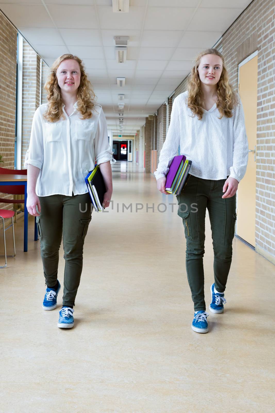 Two caucasian teenage girls walking in long school corridor carrying text books