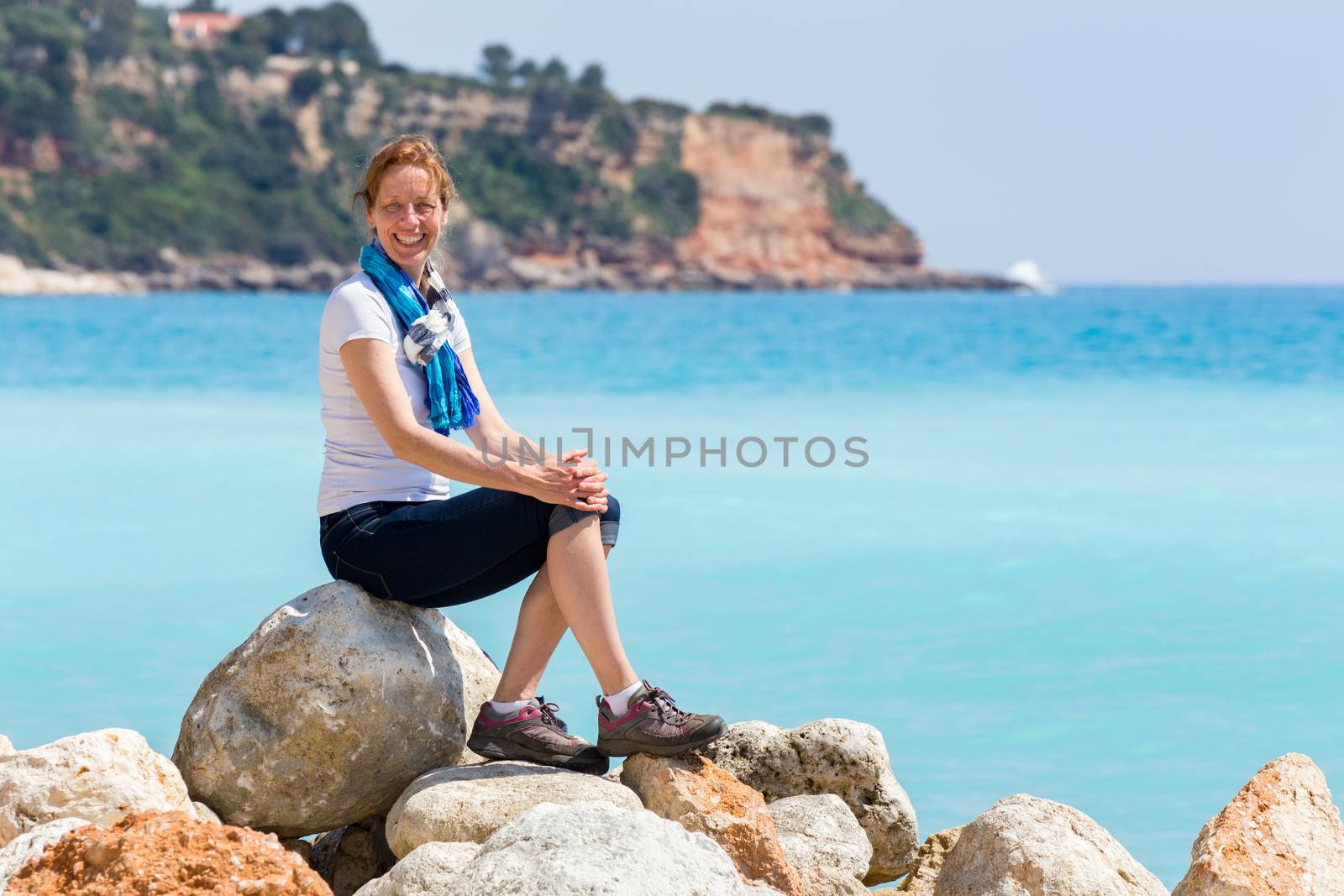 Caucasian middle aged woman as tourist sitting on rocks near blue sea enjoying vacation