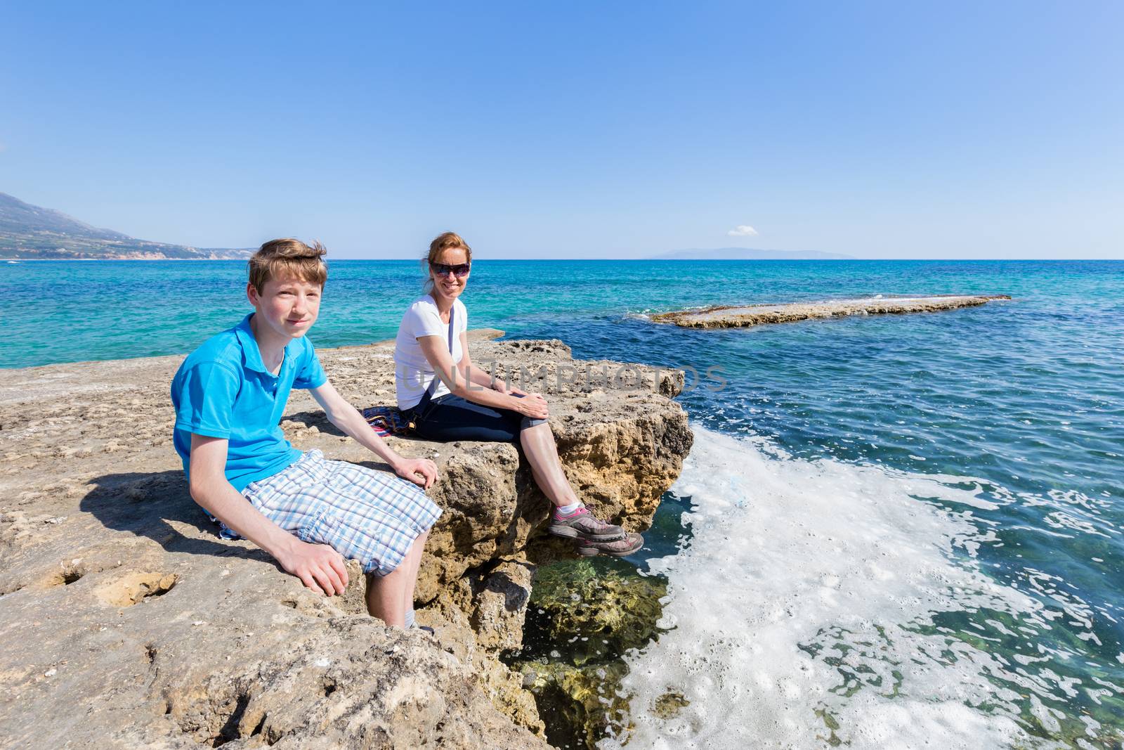 Mother and son as tourists sitting on rock at blue sea by BenSchonewille