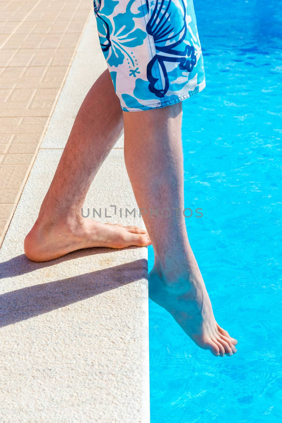 Legs of teenage boy with foot feeling water temparature in swimming pool on sunny day in summer