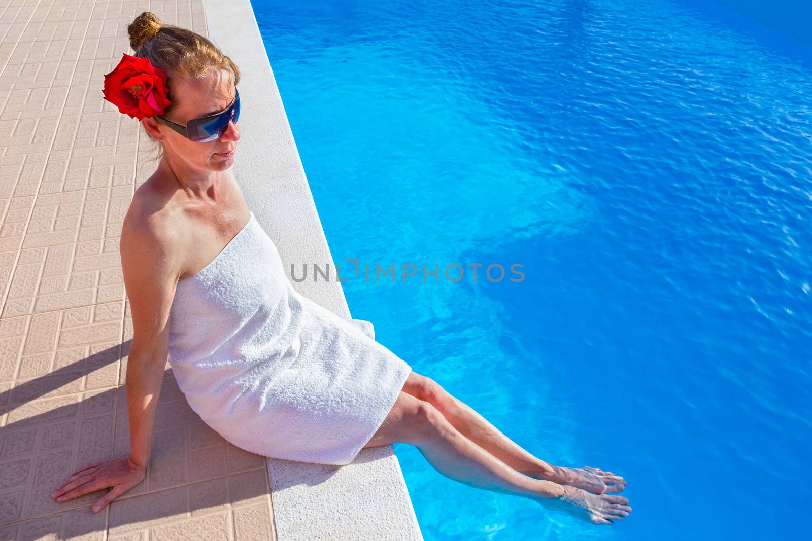 European woman wearing white towel red rose and sunglasses holding legs in blue swimming pool