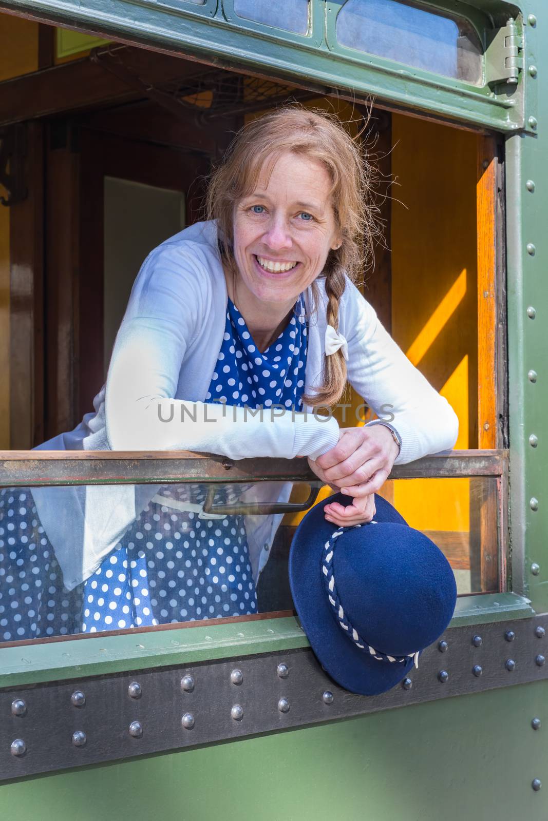 Caucasian middle aged woman dressed in blue old-fashioned clothes holding hat in train window