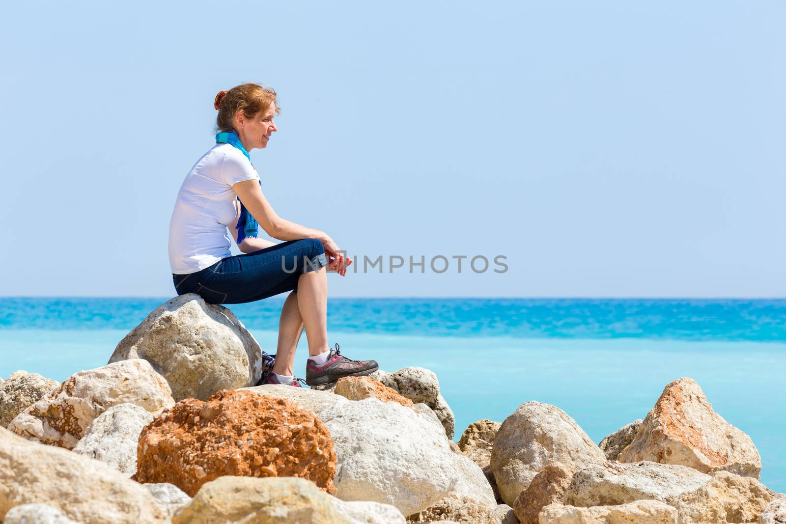 European middle aged woman sitting as tourist on rocks with blue sea horizon enjoying view