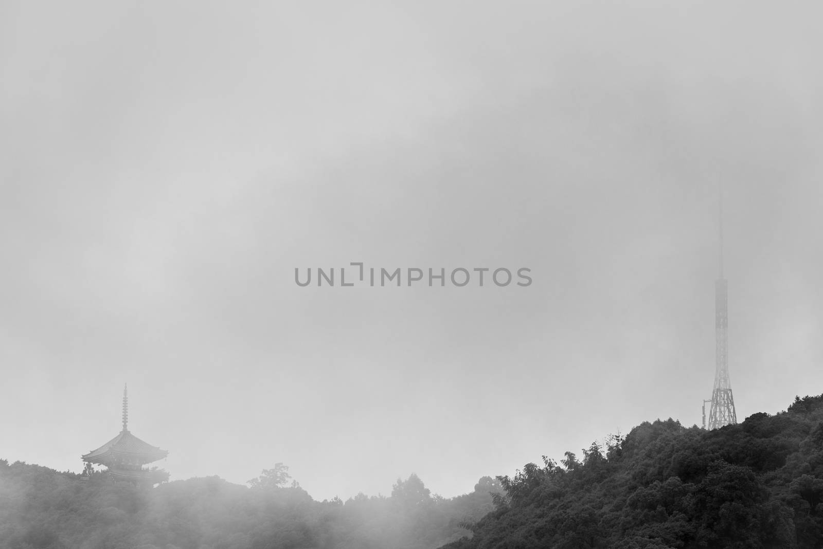 A black and white photo of an old Japanese Pagoda next to a modern radio tower on top of a mountain on a rainy, foggy day.
