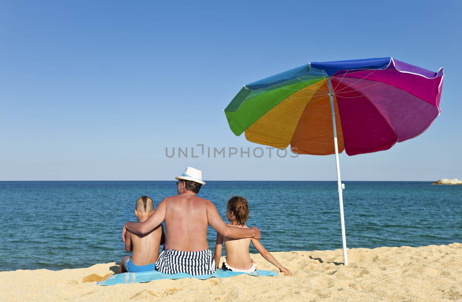 Grandfather hugging his grandson and granddaughter on a beach, looking to the see, back to the camera, colorful umbrella next to them.