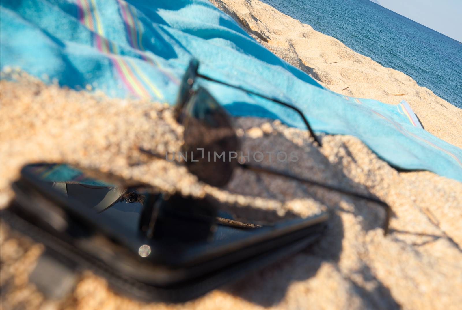 A black smart phone left buried in the send, glasses and towel on the beach with the see on focus.