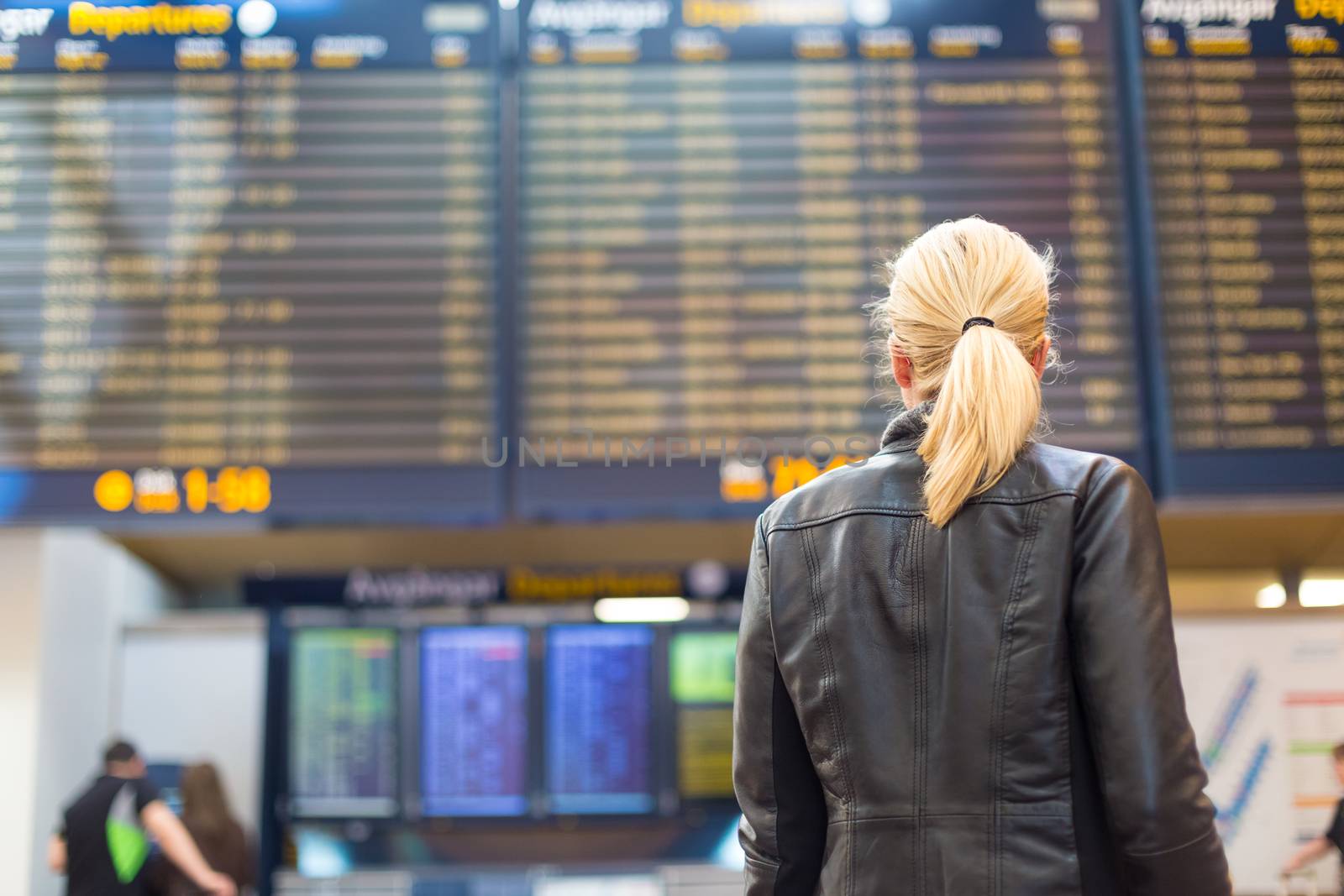 Casually dressed young stylish female traveller checking a departures board at the airport terminal hall in front of check in couters. Flight schedule display blured in the background. Focus on woman.